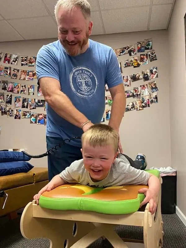 A man with a short beard and smiling is holding a young boy with a big smile, who is lying face down on a sensory Play Therapy table with a green and orange cushion. The boy is in a wooden therapy bed. Behind them, there is a wall decorated with a collage of photographs and a bench with folded blankets.