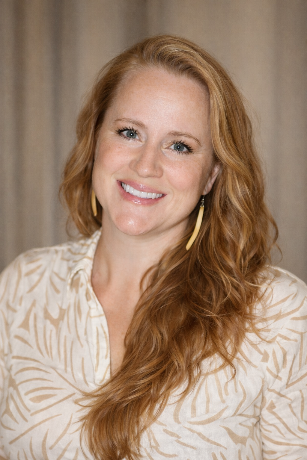 A smiling woman with long, wavy red hair, light skin, and blue eyes wearing a beige patterned blouse and gold earrings.