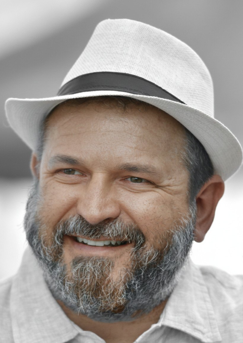 A smiling man with a gray beard and mustache wearing a white hat with a black band, dressed in a light-colored shirt.