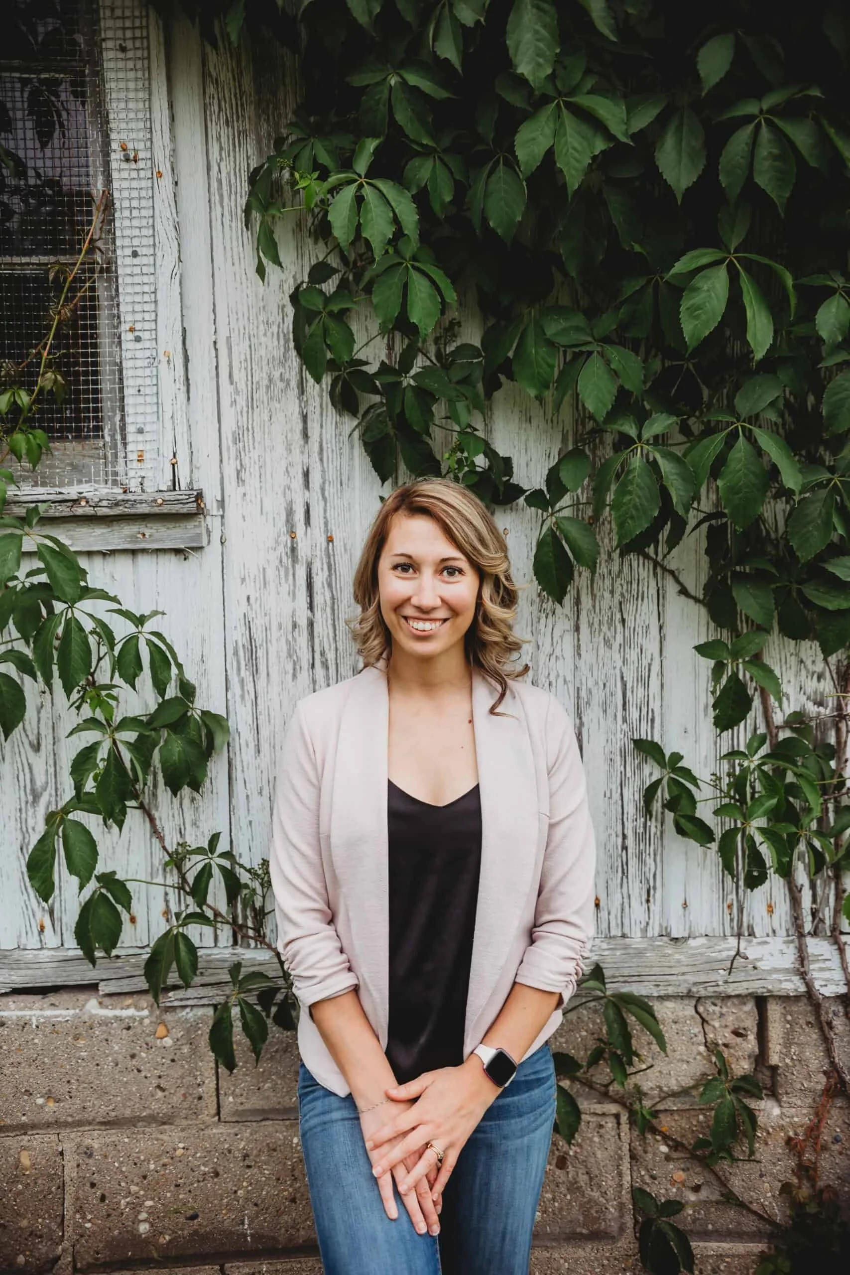 A smiling woman standing outdoors in front of a weathered white wooden wall covered with green leafy vines.