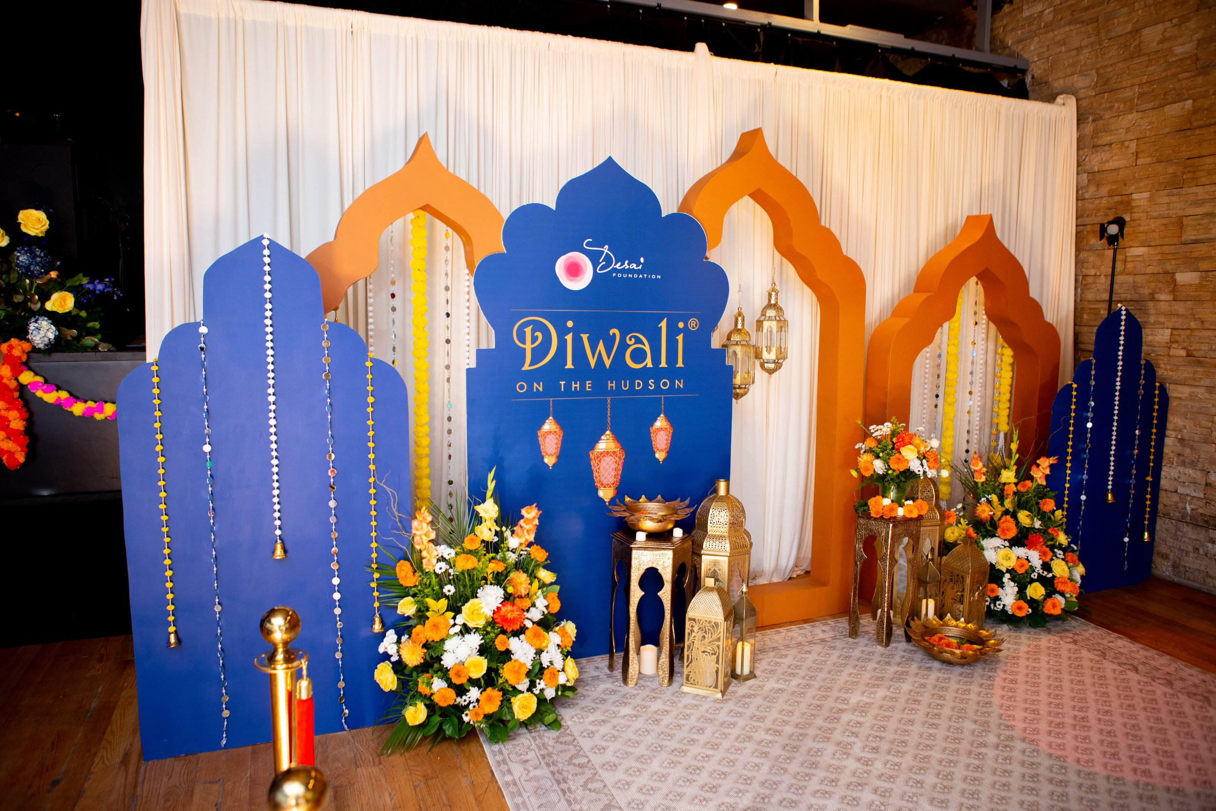 Decorative setup with blue and orange arch shapes, floral arrangements, lanterns, and a sign that reads 'Diwali on the Hudson,' indicating a Diwali celebration event.