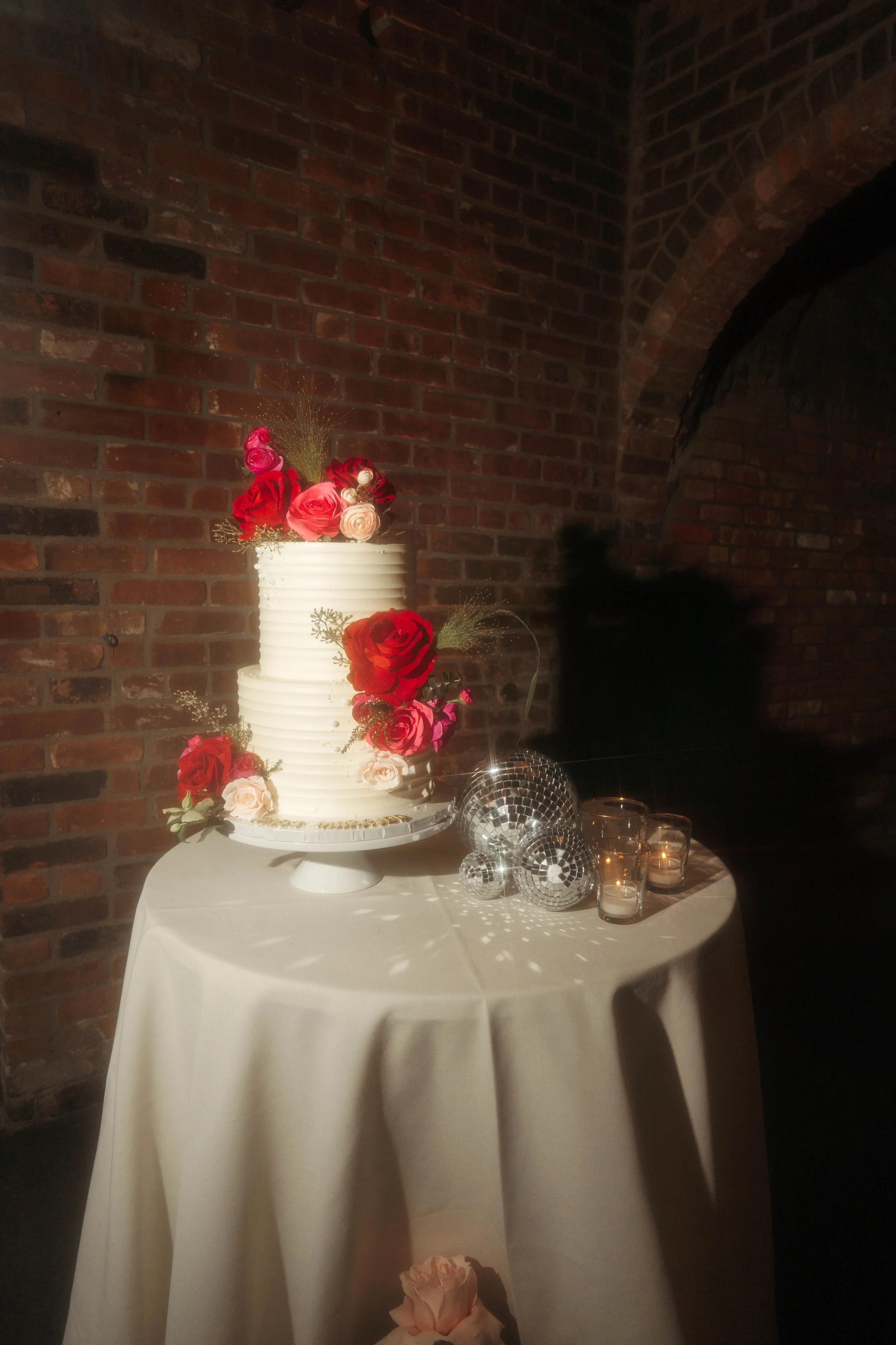 A multi-tiered white wedding cake decorated with pink, red, and peach roses, set on a white table with a brick wall background. Next to the cake are glass disco ball decorations and small candles.