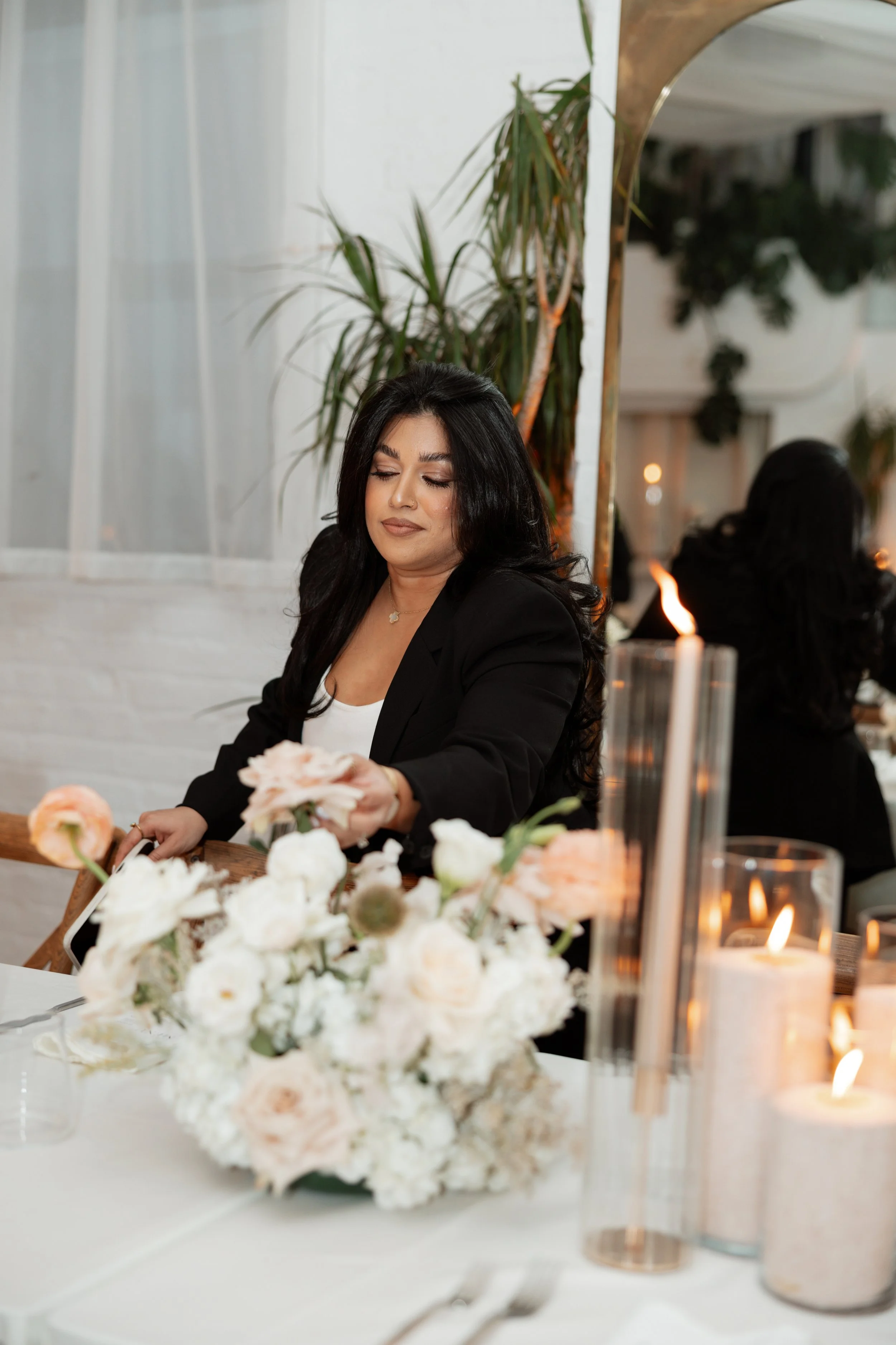 A woman with long dark hair wearing a black blazer and white top is arranging flowers on a table decorated with candles and floral arrangements in a bright room with green plants and a mirrored wall.