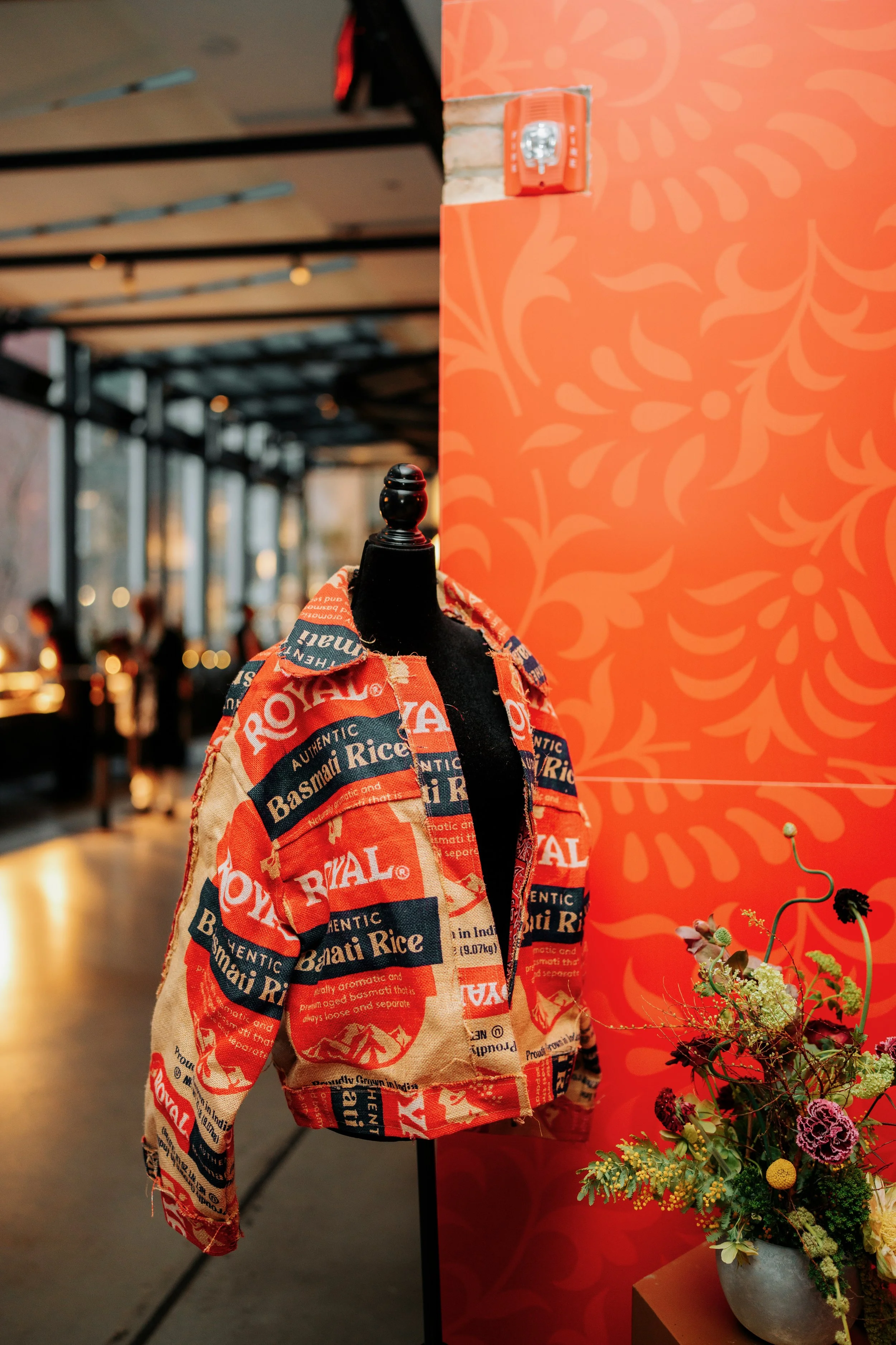 A display with a bag made from rice sacks hanging on a black mannequin and a bouquet of flowers in a gray pot on a small table next to a bright orange decorative wall with a floral pattern.