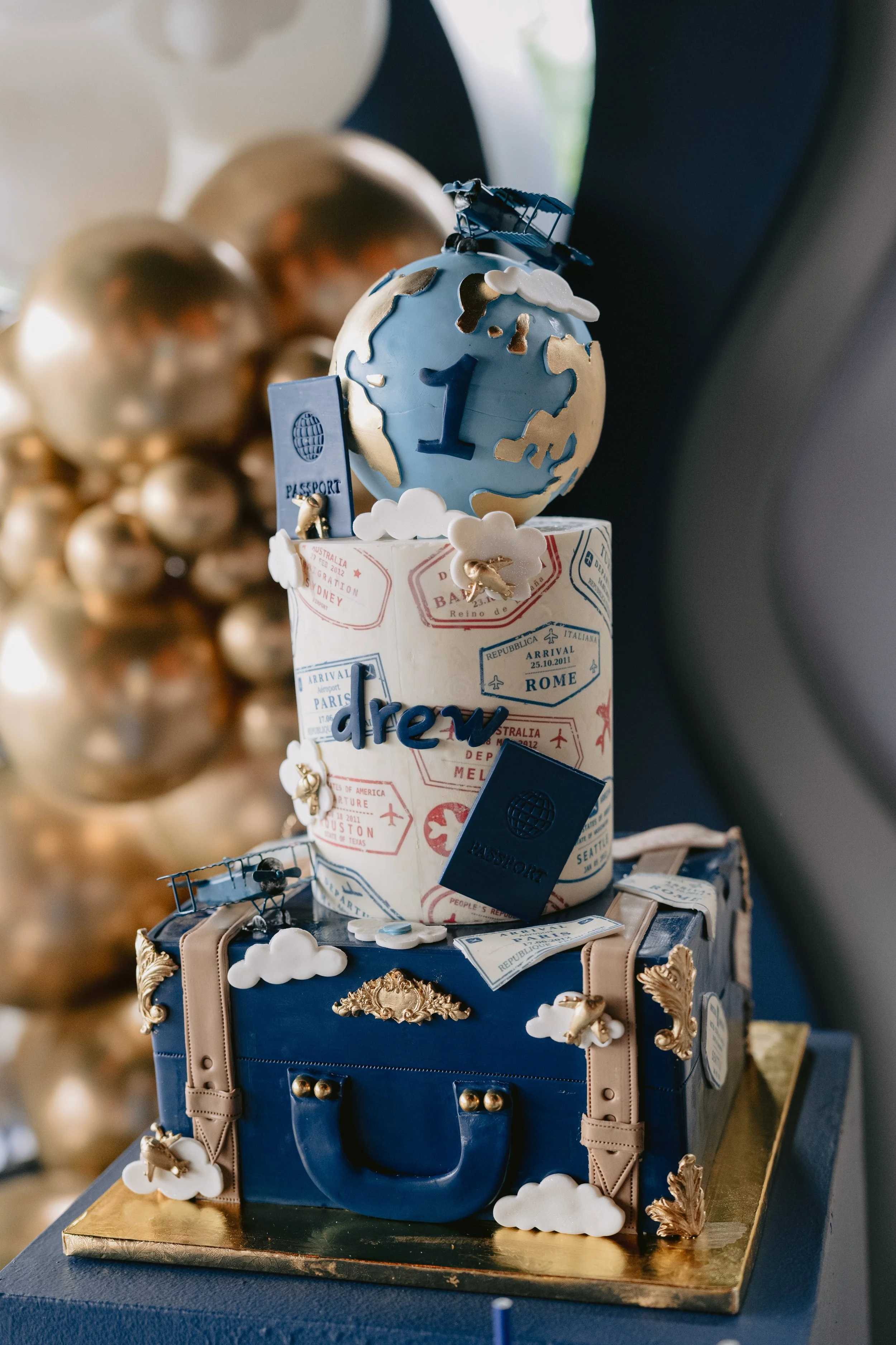 A suitcase-shaped cake decorated with golden leaves and clouds, featuring travel-themed elements like a miniature airplane and passport, with a globe cake on top displaying a large blue '1' and an airplane figure, sitting on a gold-colored base.