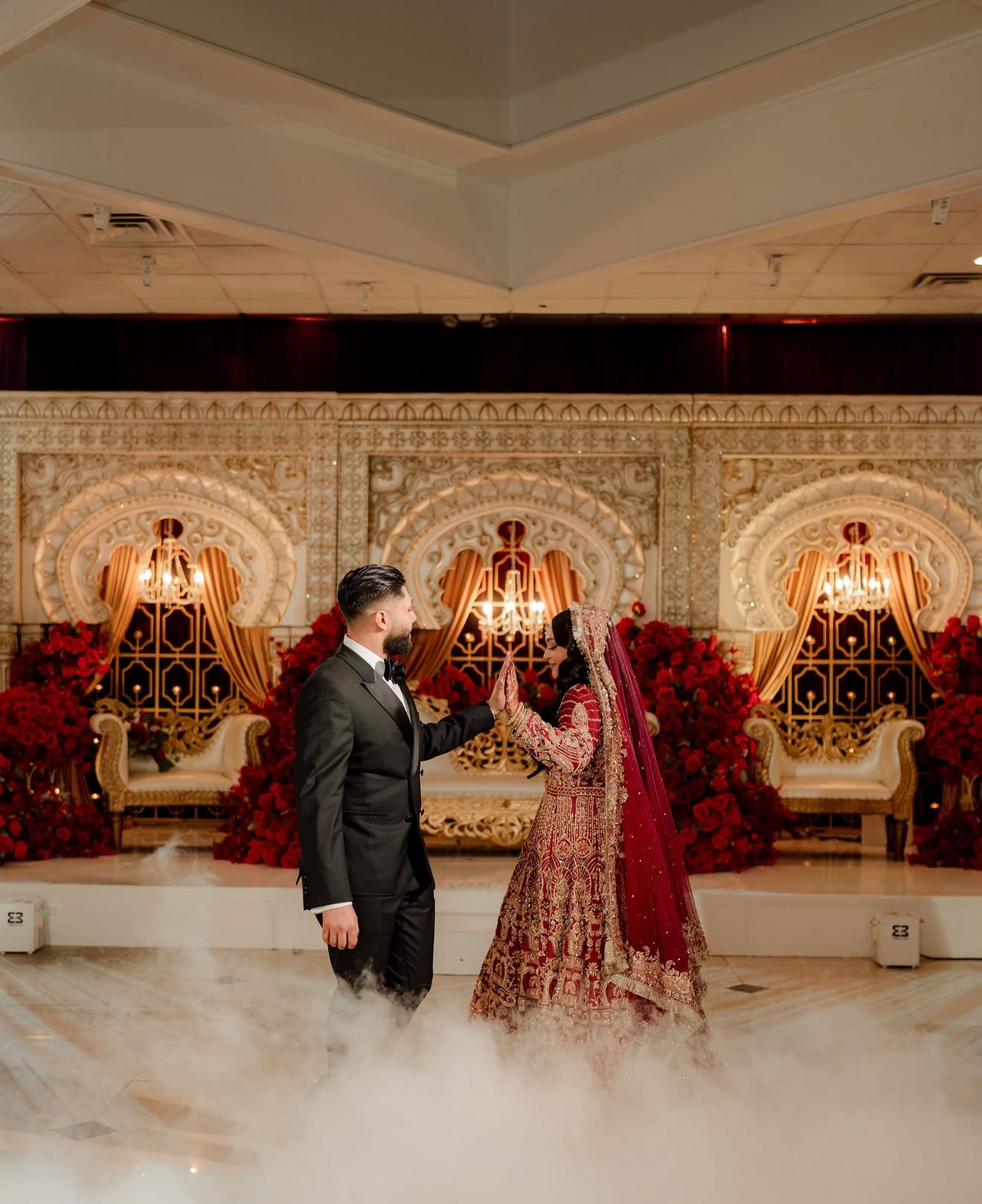 A bride and groom dancing during a wedding ceremony in a grand hall decorated with red roses, ornate decor, and chandeliers.