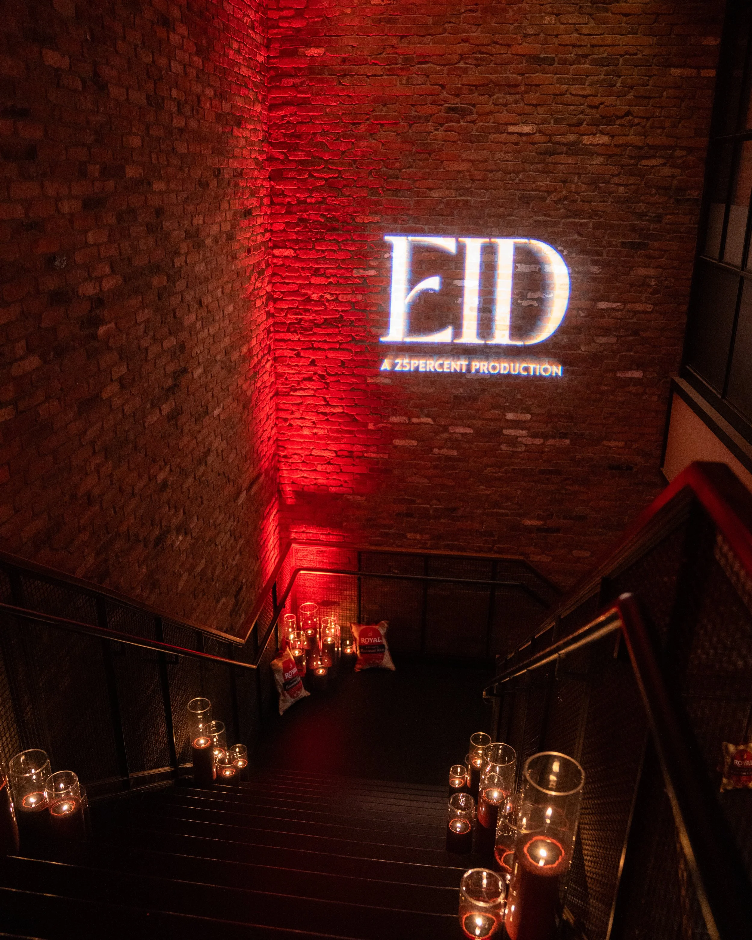 A brick wall with a neon sign reading 'EID' and smaller text underneath stating 'A 25percent production.' There are candles in glass holders along a staircase leading downward, with some bags of Royal brand pet food at the bottom of the stairs. The s