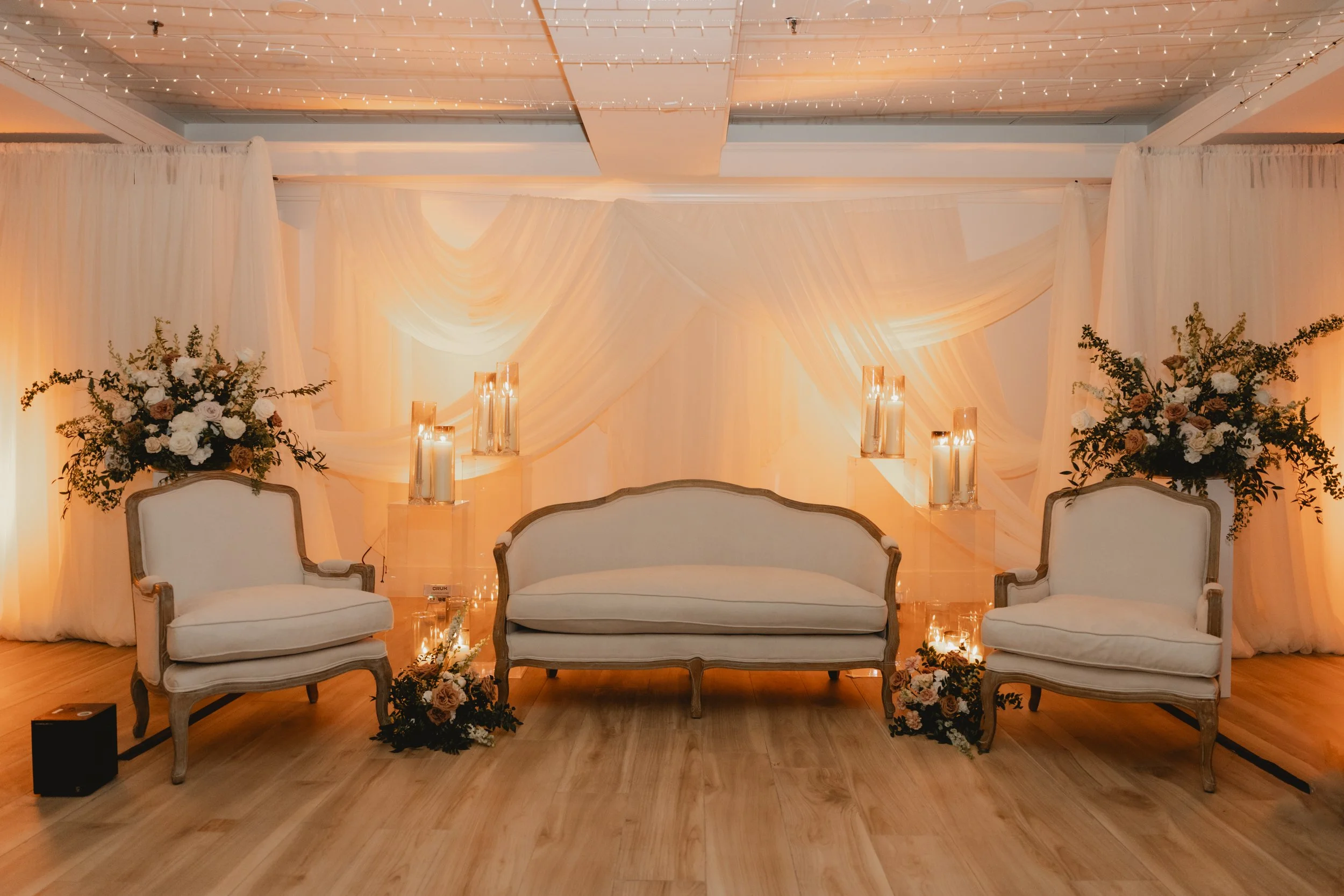 Elegant event space with white vintage-style sofas, floral arrangements, candlesticks, and draped white fabric backdrop with string lights.