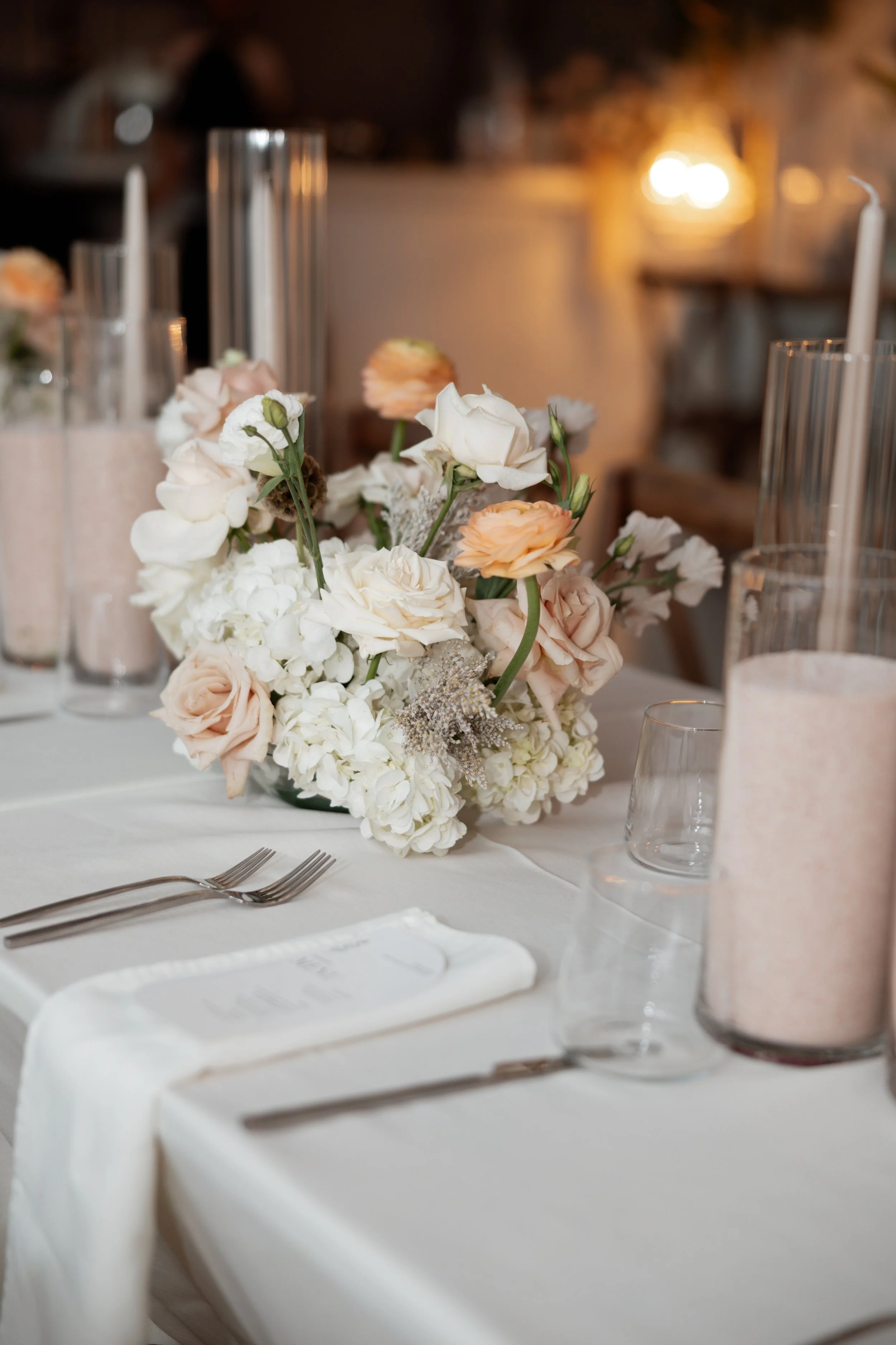 A table set for a formal event with a centerpiece made of white and blush flowers, surrounded by glassware, candles, and cutlery, with a blurred background of warm lighting.