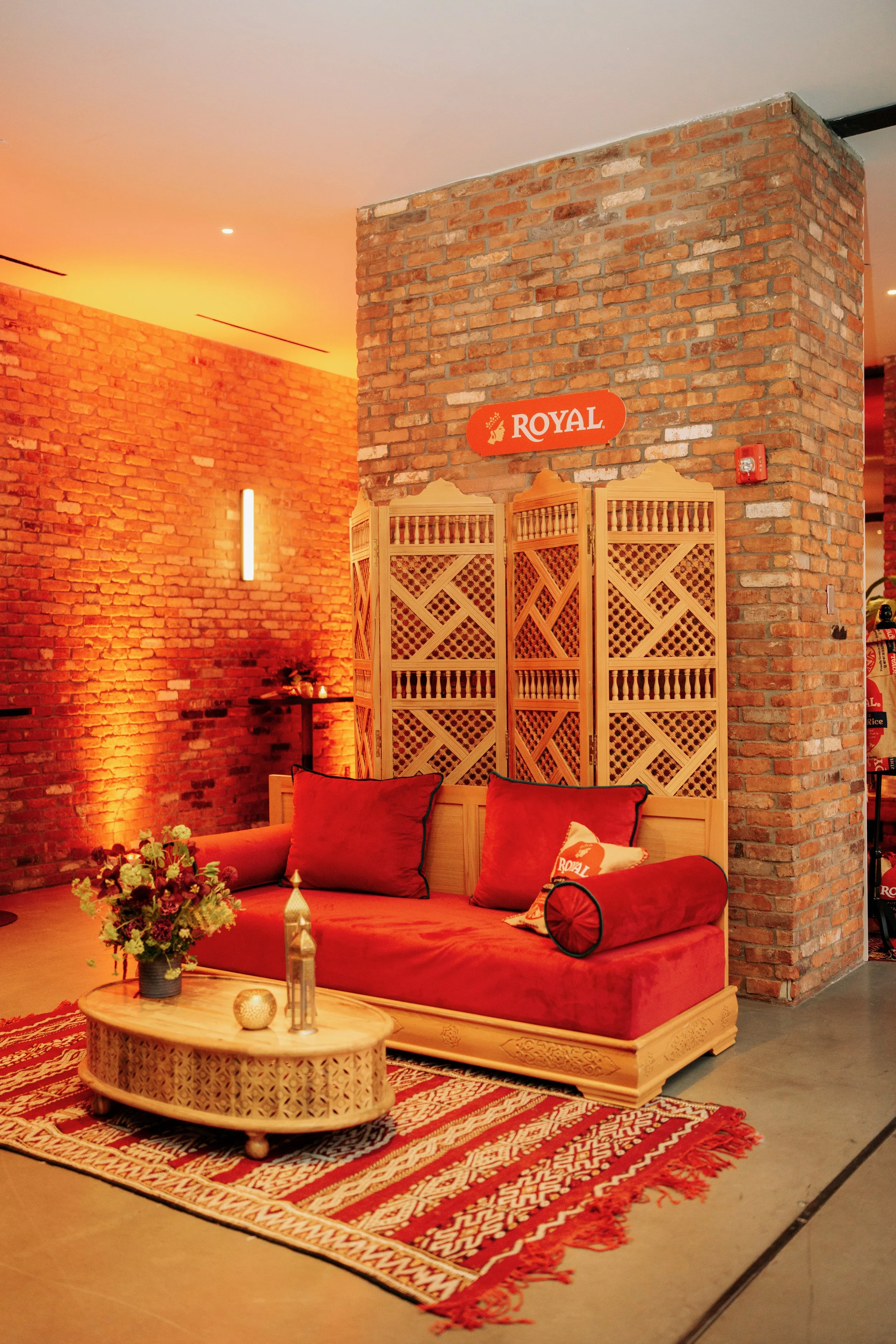A cozy living room corner with a red velvet sofa, red pillows, and a wooden coffee table with a flower arrangement and decorative items. Exposed brick walls and warm lighting create a welcoming atmosphere.
