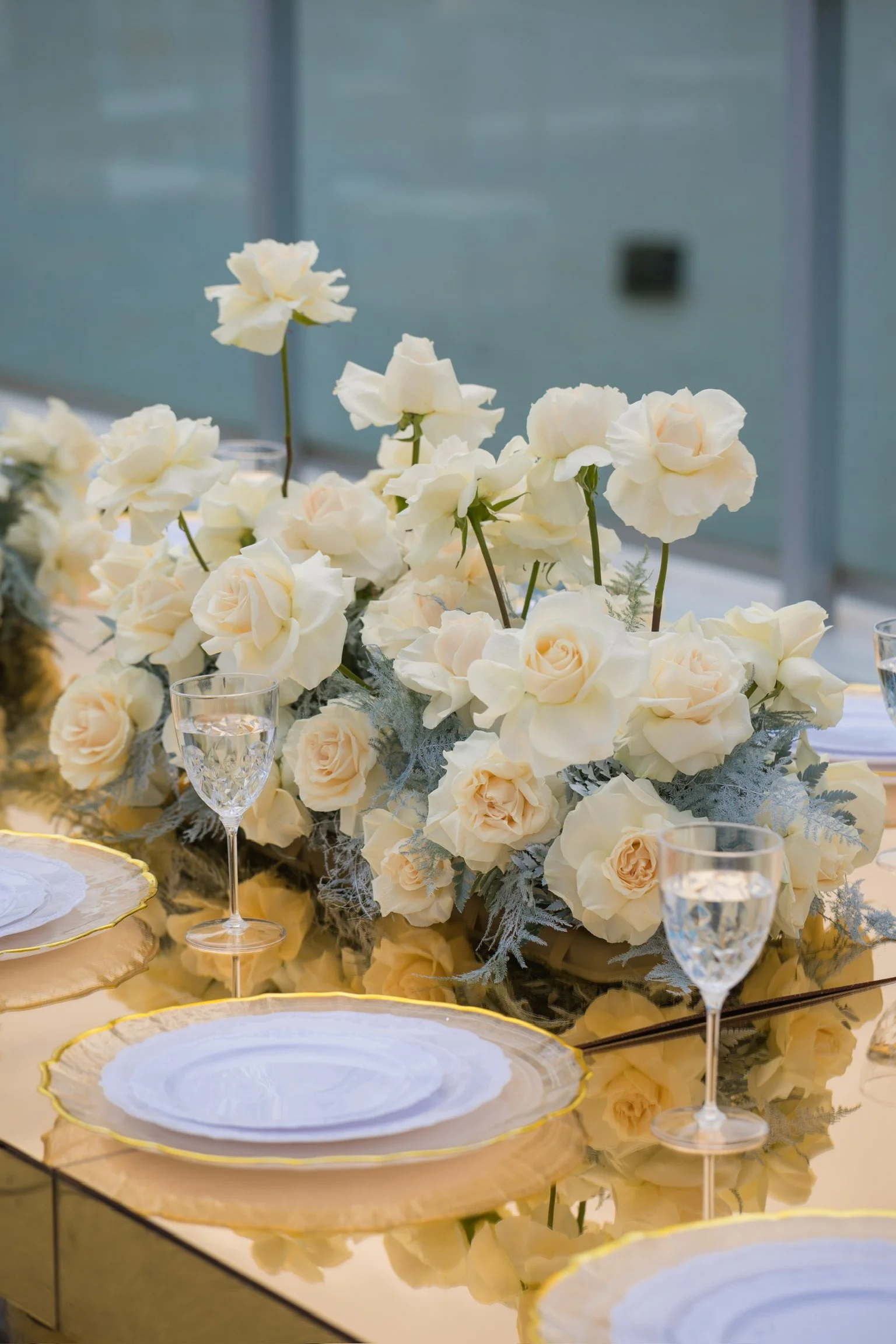 Elegant table setting with white roses centerpiece, glassware, and gold-rimmed plates on a reflective gold table at a formal event.