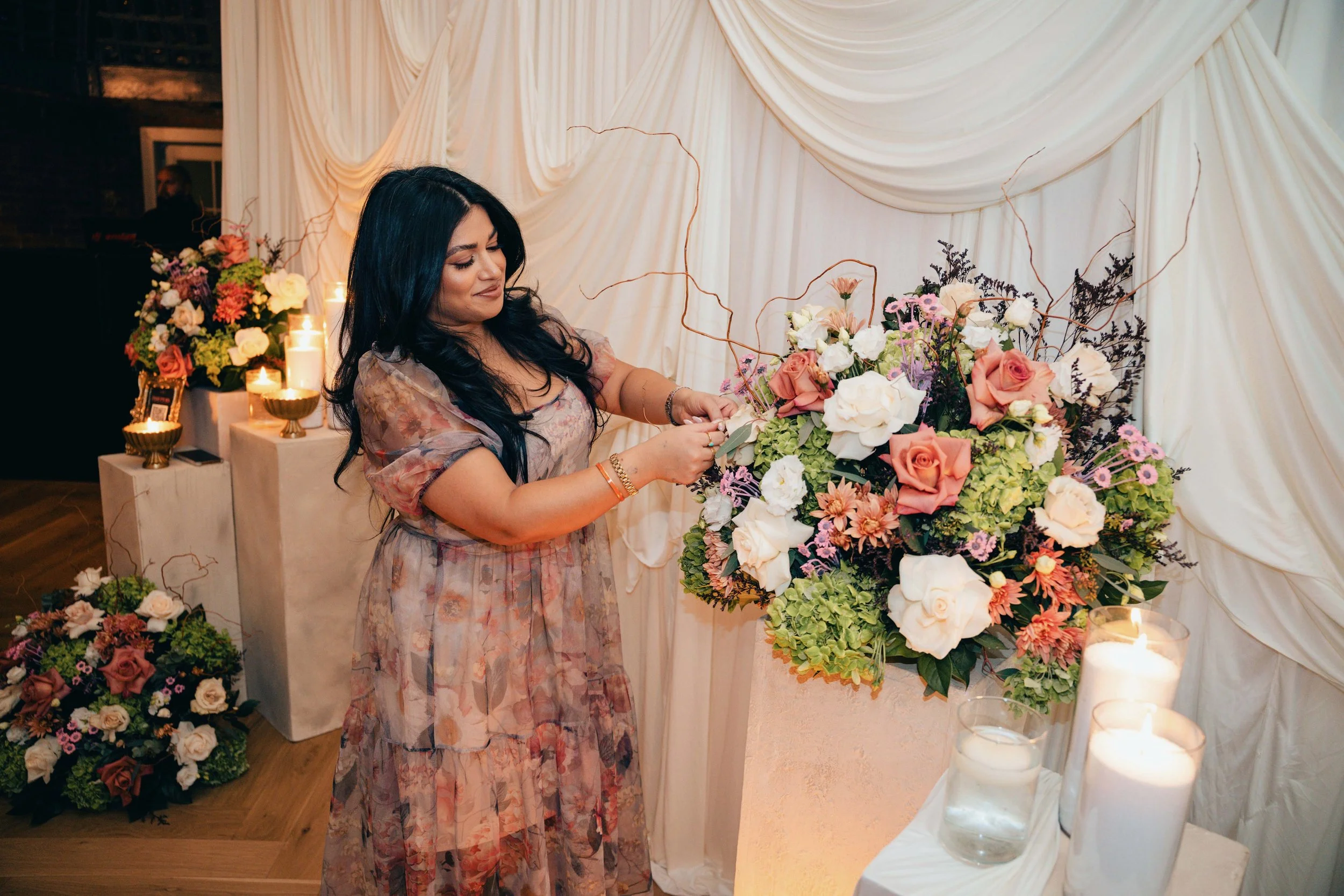 A woman with long dark hair, wearing a floral dress, arranging a large bouquet of flowers at a wedding or celebration decor setting. Behind her, a backdrop of white draped fabric, with candles and smaller floral arrangements on tables.