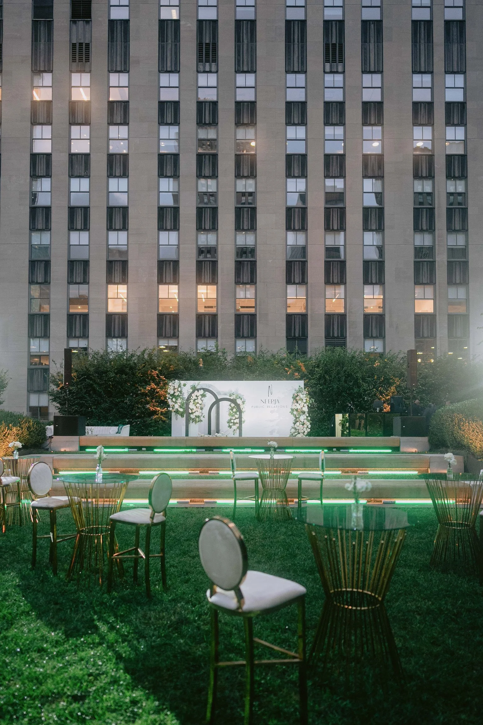 An outdoor event space at dusk with tables and chairs on a grassy area, illuminated with green accent lighting, in front of a tall building with lit windows.