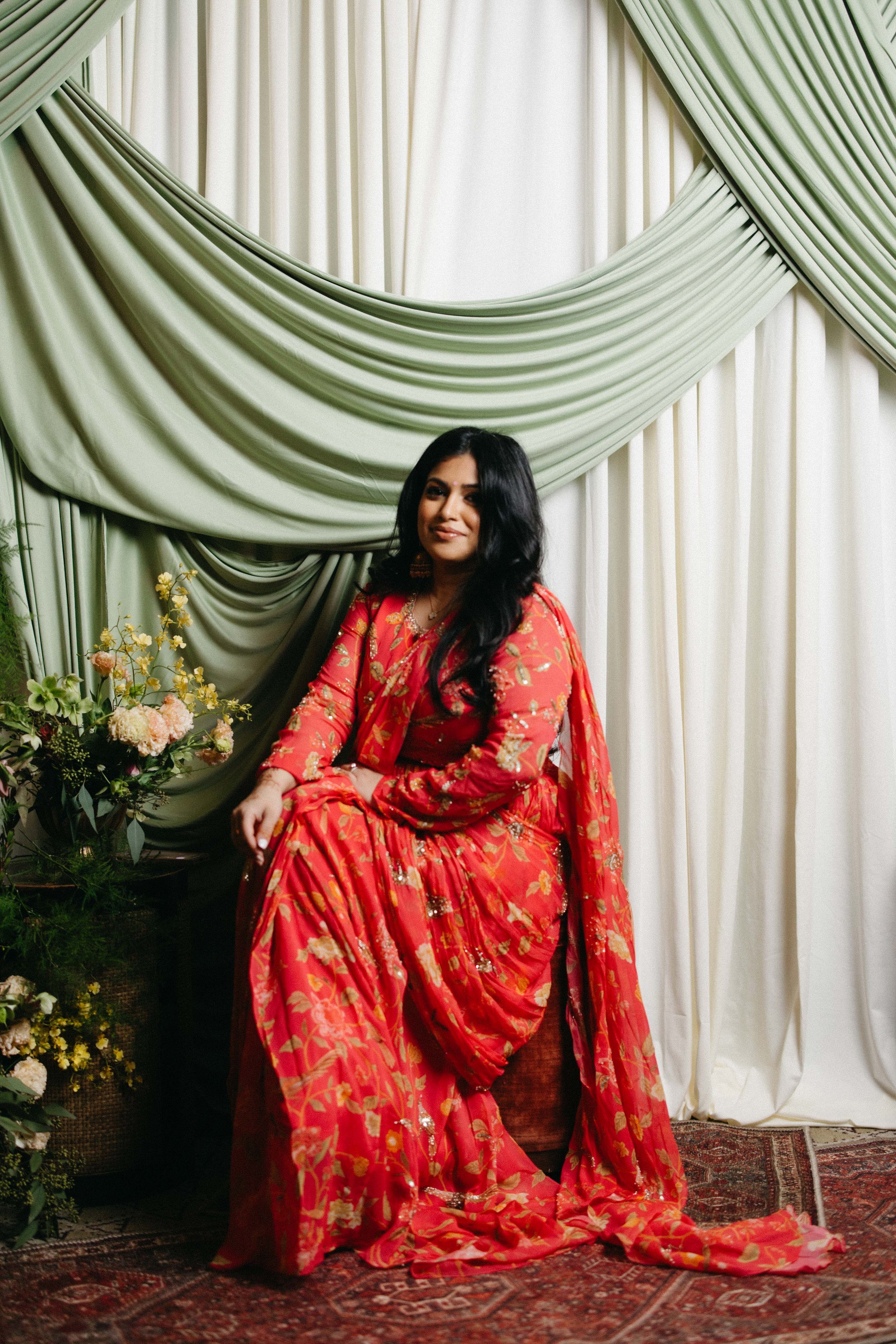 A woman dressed in a traditional red and gold sari sitting on a patterned rug in front of green and white draped curtains, with flowers to her left.