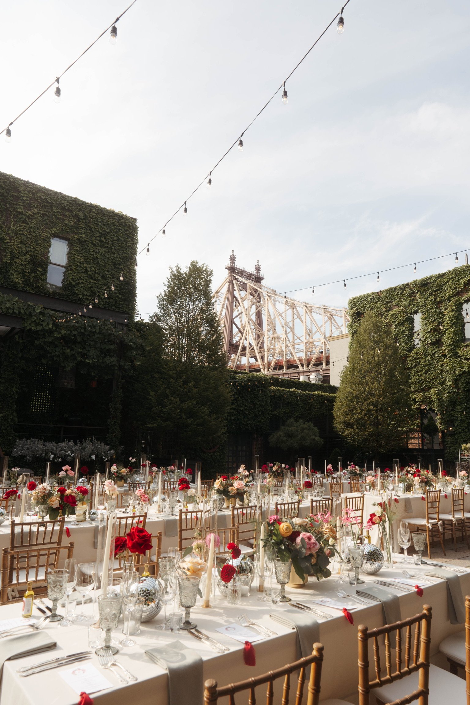 An outdoor wedding reception with tables decorated with flowers and candles, set up in an urban open-air space with string lights overhead, tall trees, and a view of a bridge in the background.