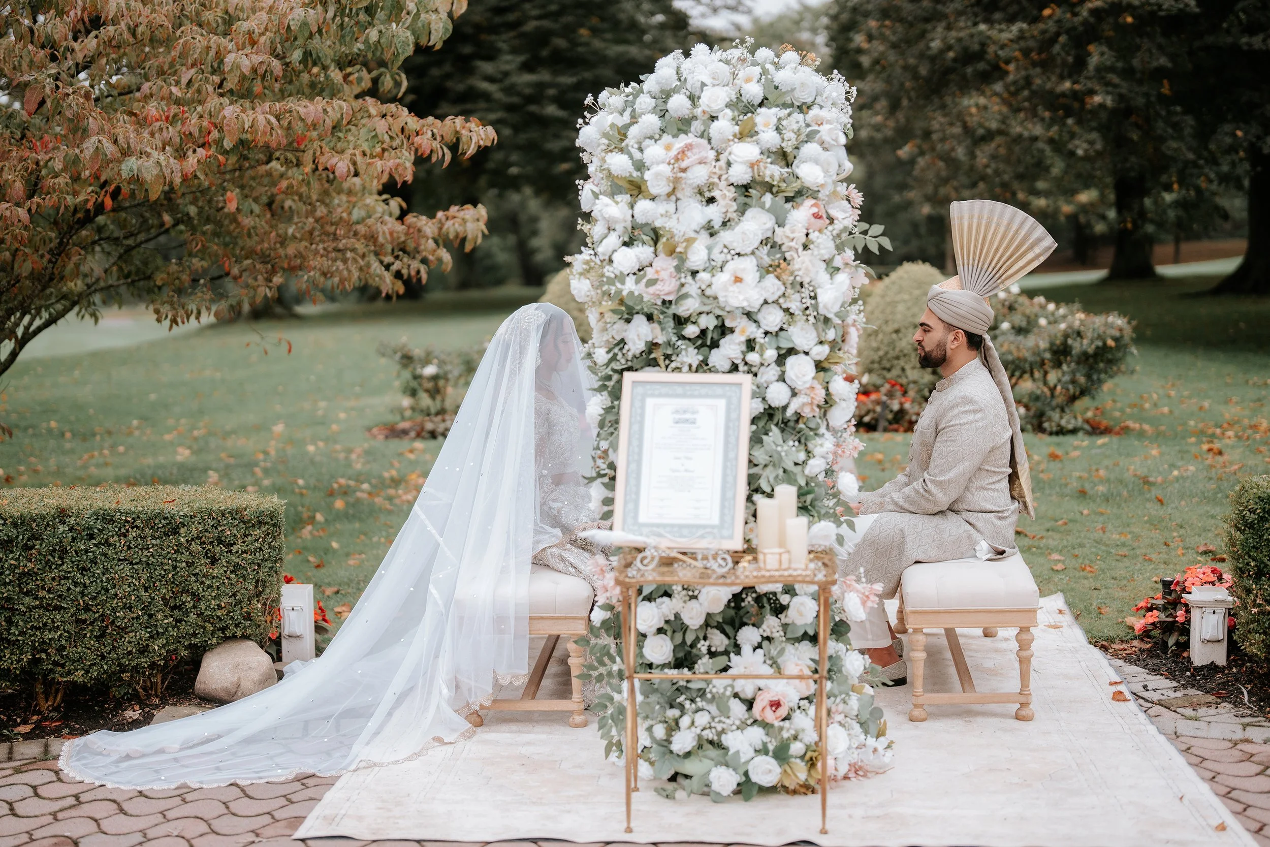 A wedding ceremony outdoors with a bride and groom kneeling in front of a tall floral arrangement. The bride is wearing a white wedding dress with a veil, and the groom is dressed in traditional Indian attire with a turban. There are candles and a fr