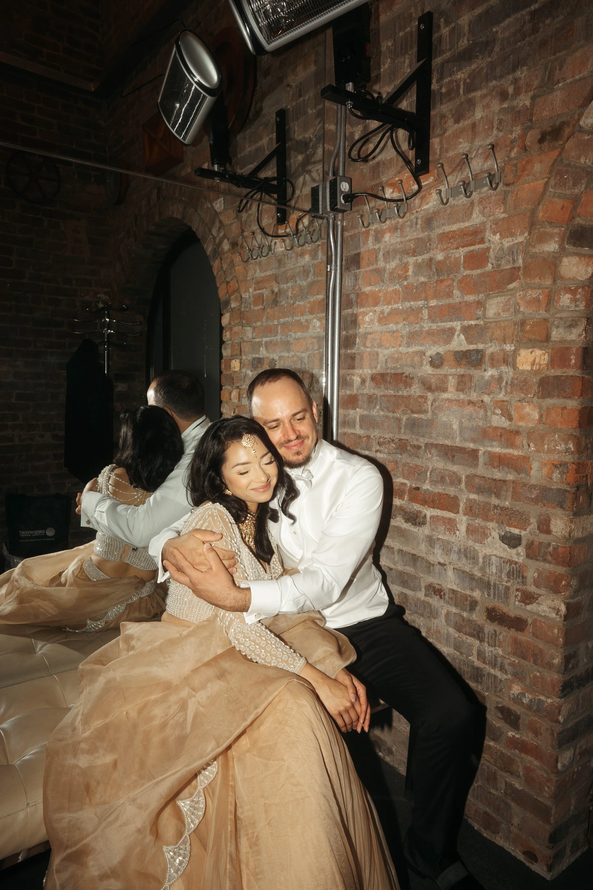 A group of people embracing closely and smiling in a cozy indoor setting with brick walls and warm lighting.