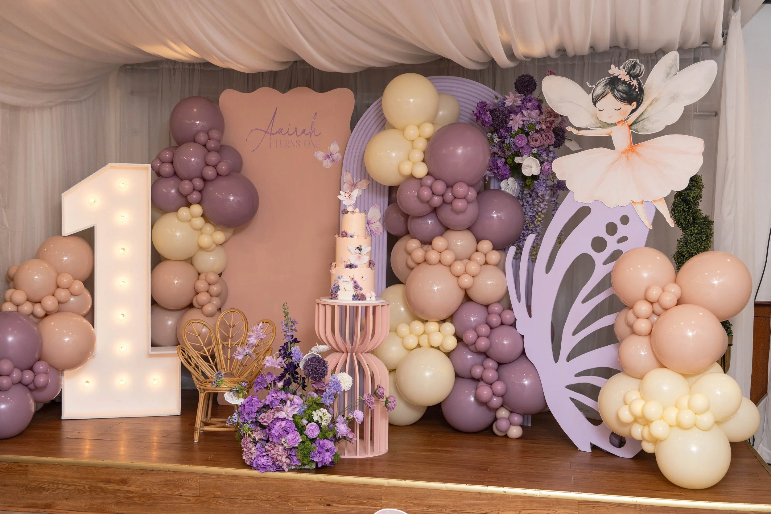 Decorative birthday backdrop with purple, cream, and pink balloons, flowers, and a cutout of a fairy girl in a pink tutu for a first birthday celebration.