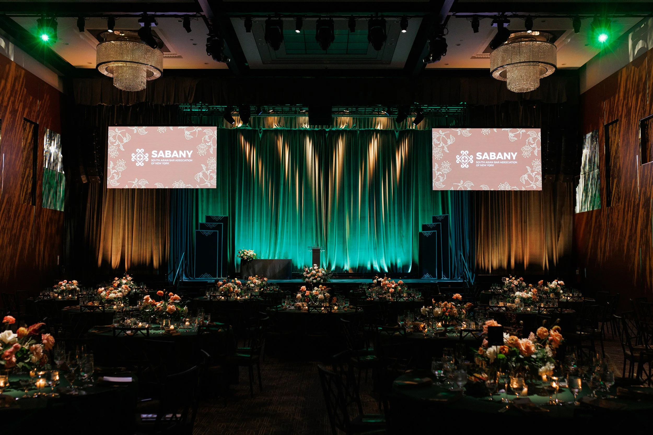 A large event hall decorated for a formal gathering with floral arrangements and a stage, displaying screens with the logo of the South Asian Bar Association of New York.