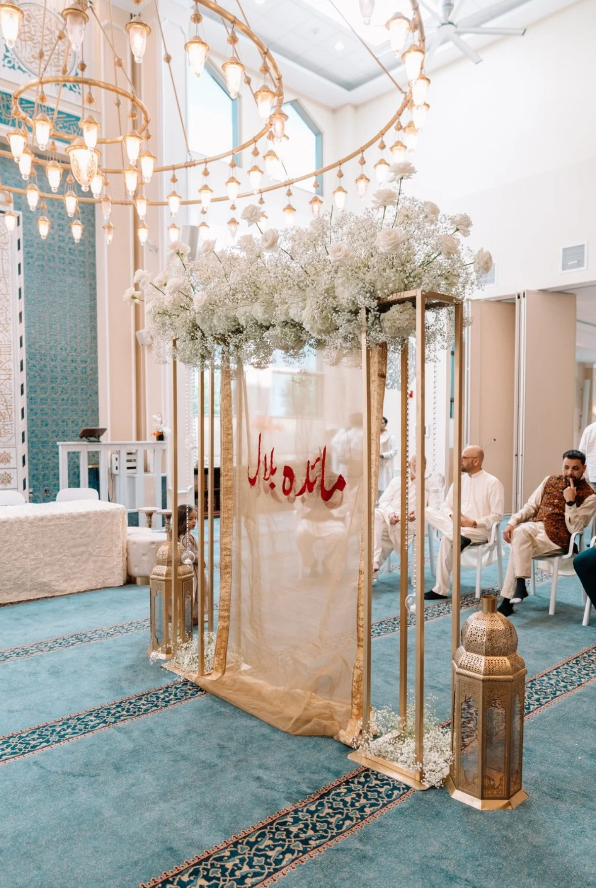 A decorated wedding or event space with a large floral arrangement of white flowers and greenery atop a gold frame. The space includes ornate lanterns, a blue carpeted floor, and white seating. A beige fabric banner with red Arabic script hangs on th