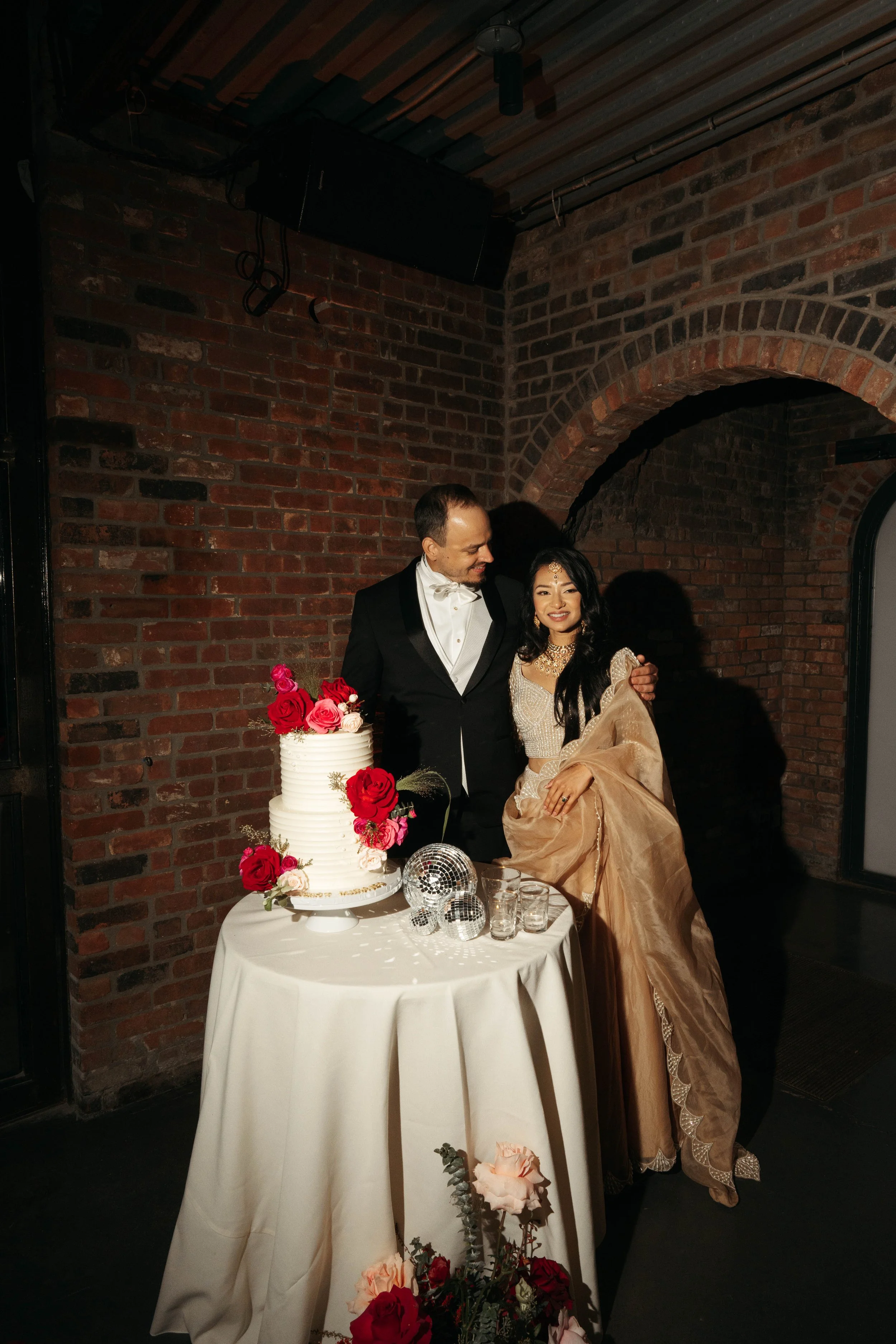 A bride and groom standing next to a wedding cake decorated with red and pink roses, with a brick wall background and a table with flowers and decorative items in the foreground.