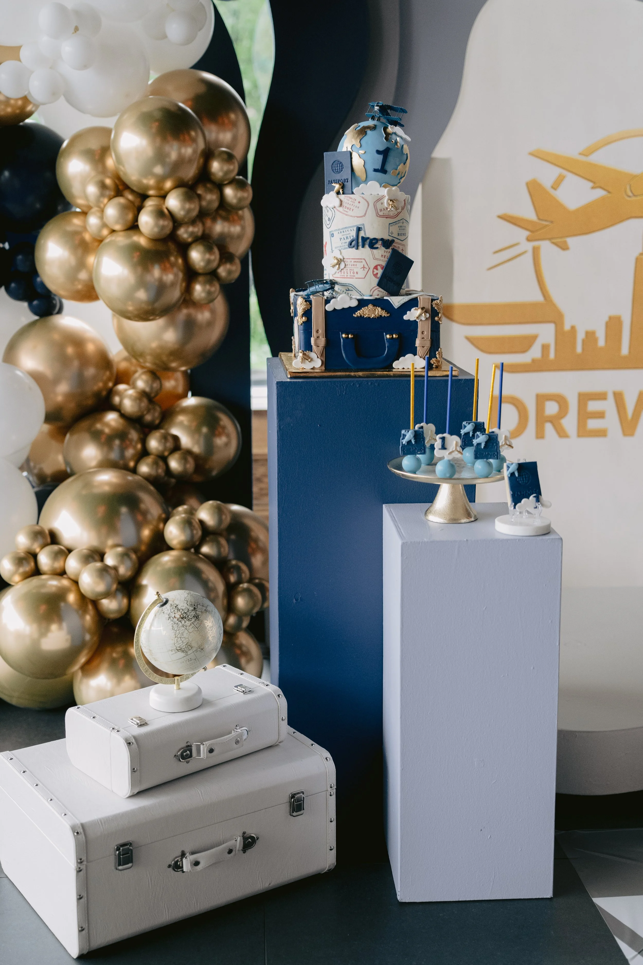 Decorative globe on a white suitcase, surrounded by a balloon display with gold, black, and white balloons, positioned next to a blue and white themed dessert table with travel-inspired decorations and a large airplane graphic on the wall.