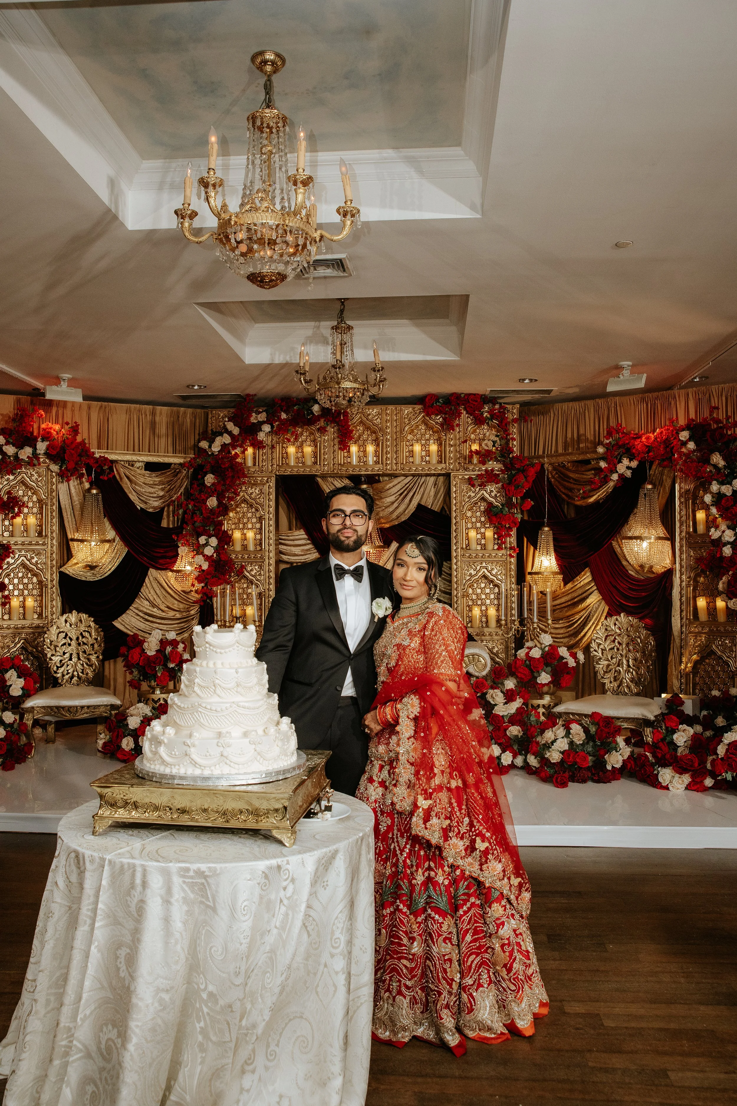 A bride and groom standing together at their wedding reception in front of a decorated backdrop with red and white flowers, candles, and drapery. The groom is wearing a black tuxedo, and the bride is wearing a traditional red and gold bridal dress. A