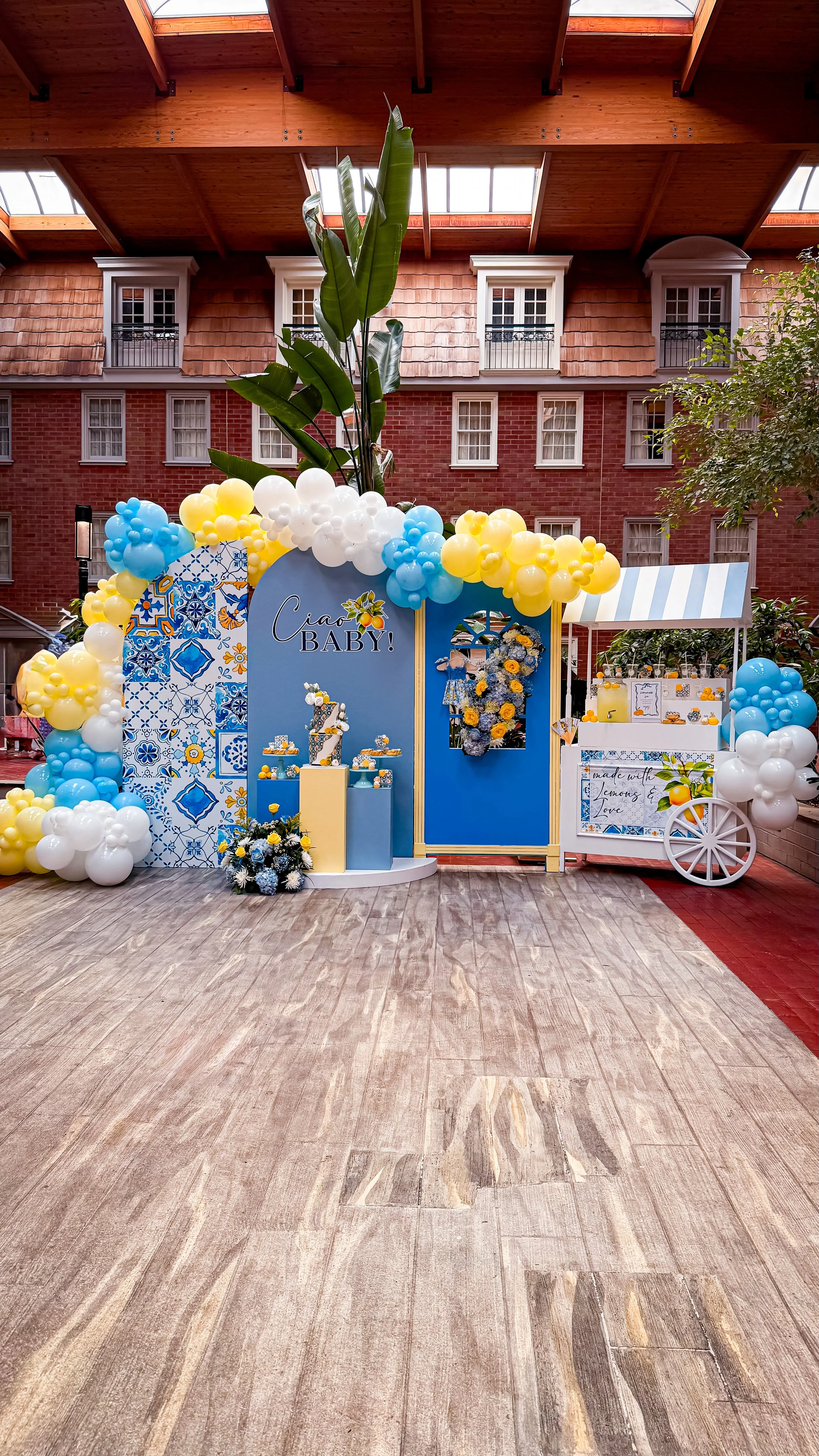 Decorative celebration setup for a baby shower or arrival, featuring blue, yellow, and white balloons, a blue backdrop with the words 'Ciao BABY!', floral arrangements, and a small cart with drinks and decorations, under a wooden ceiling with skyligh