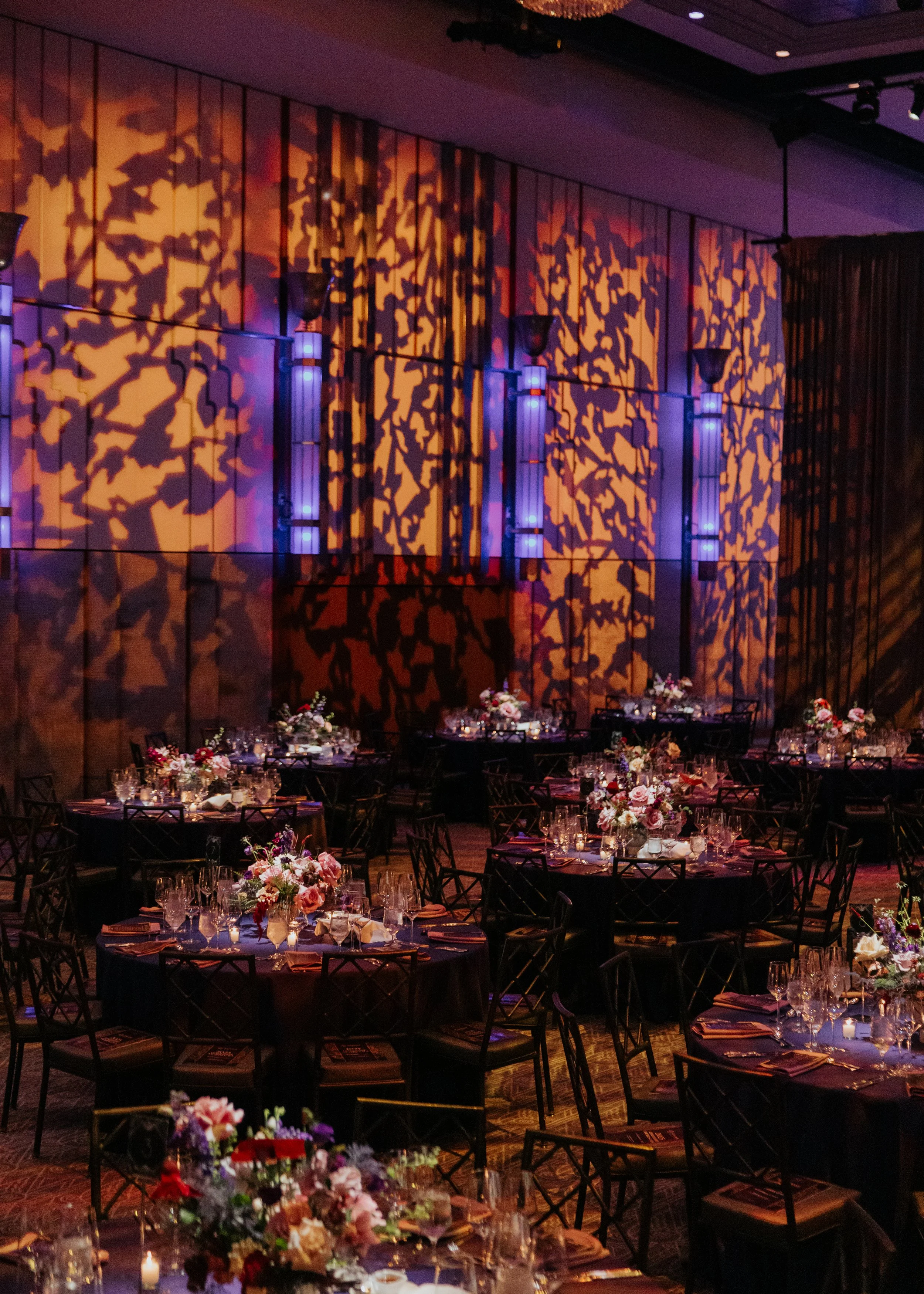 Elegant banquet hall decorated for an event with round tables covered in dark tablecloths, floral centerpieces, and neatly arranged glassware and table settings. The wall features dramatic orange and purple lighting projections creating a leaf patter