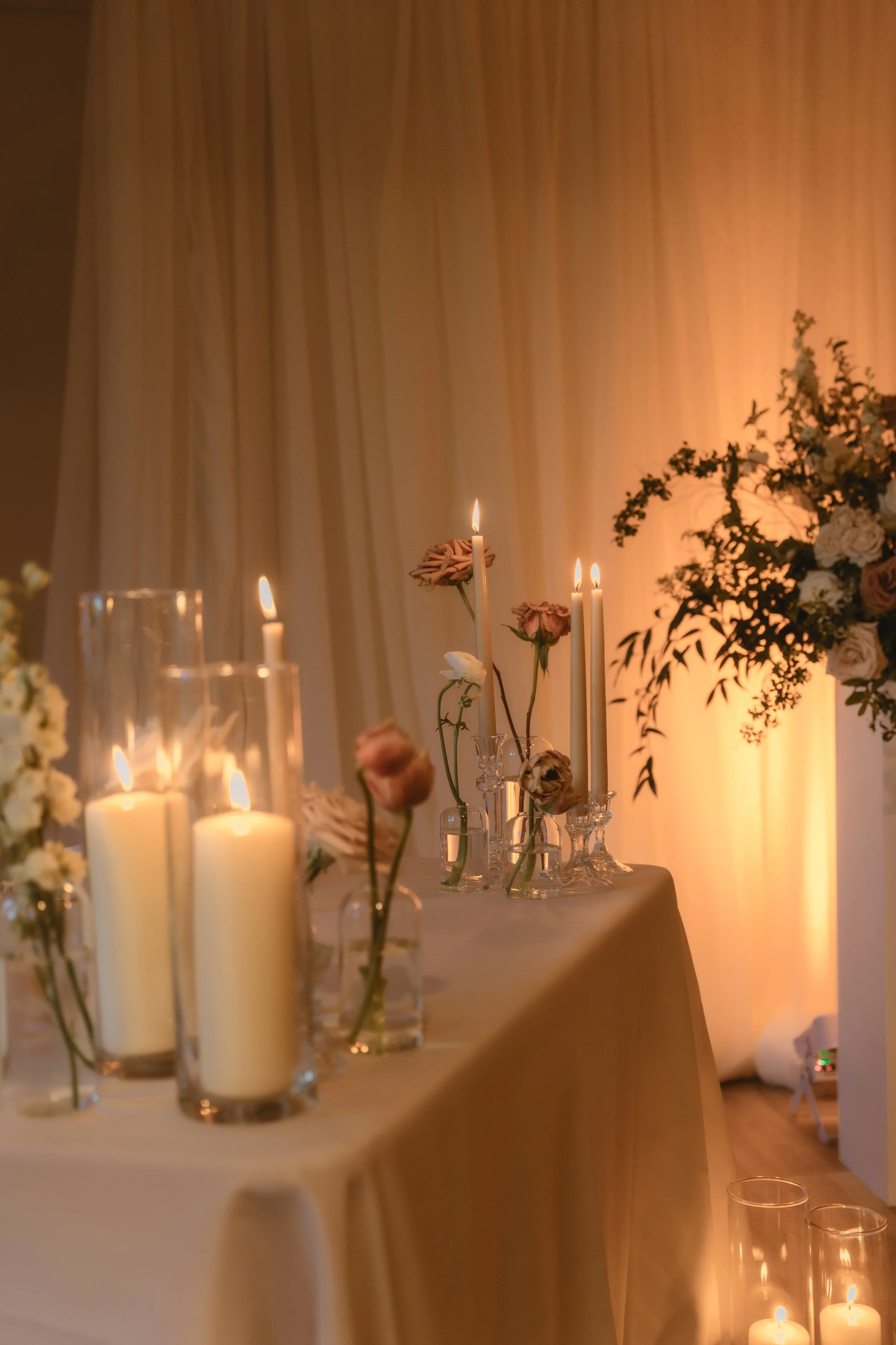 A table decorated for a celebration with candles and floral arrangements. The background features soft, warm lighting and cream-colored drapes.