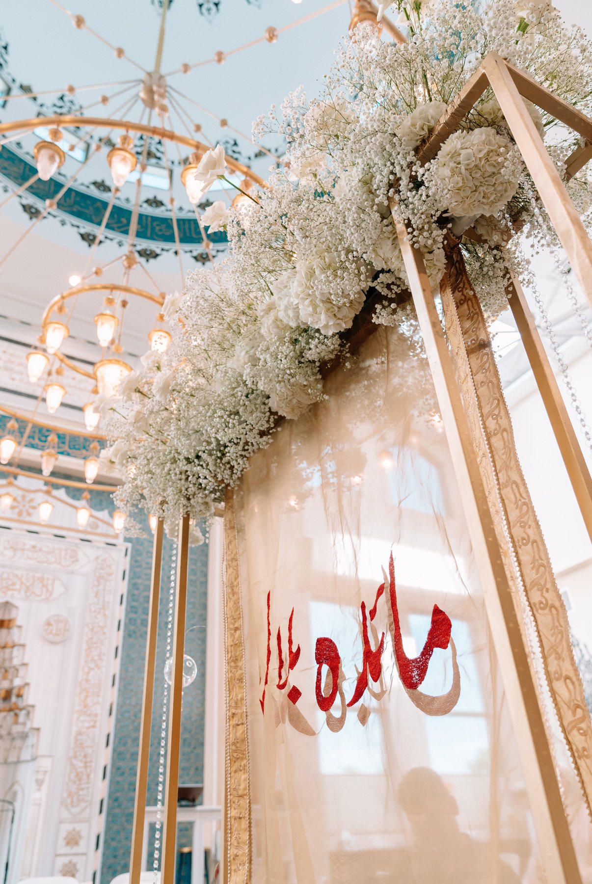 Decorative floral arrangement with white flowers on a golden stand, with a banner containing red and gold lettering, set in an elegant room with chandeliers and intricate ceiling details.