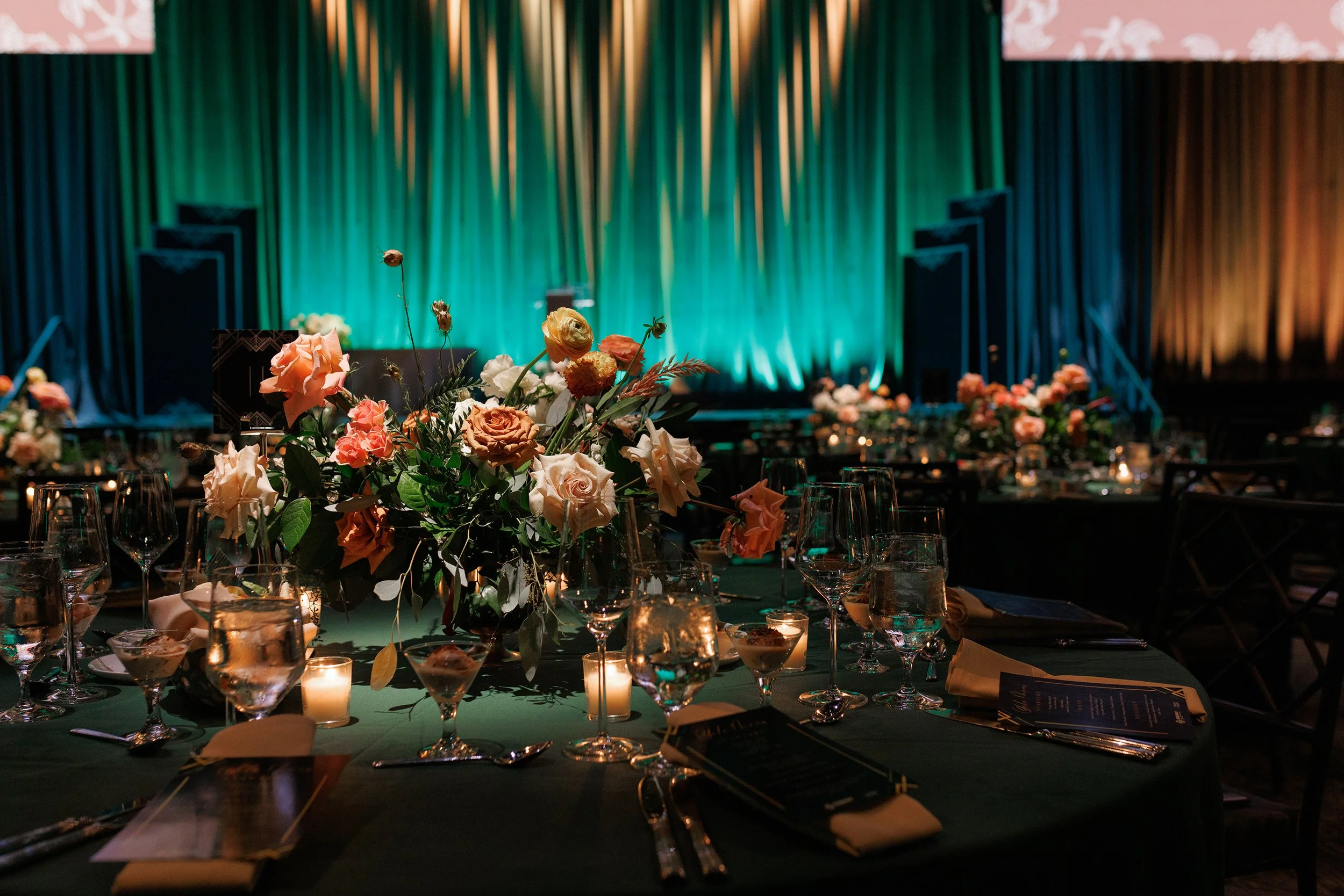 A decorated banquet table with glassware, candles, and floral centerpieces, set for a formal event in a dimly lit room with a stage and teal curtains in the background.