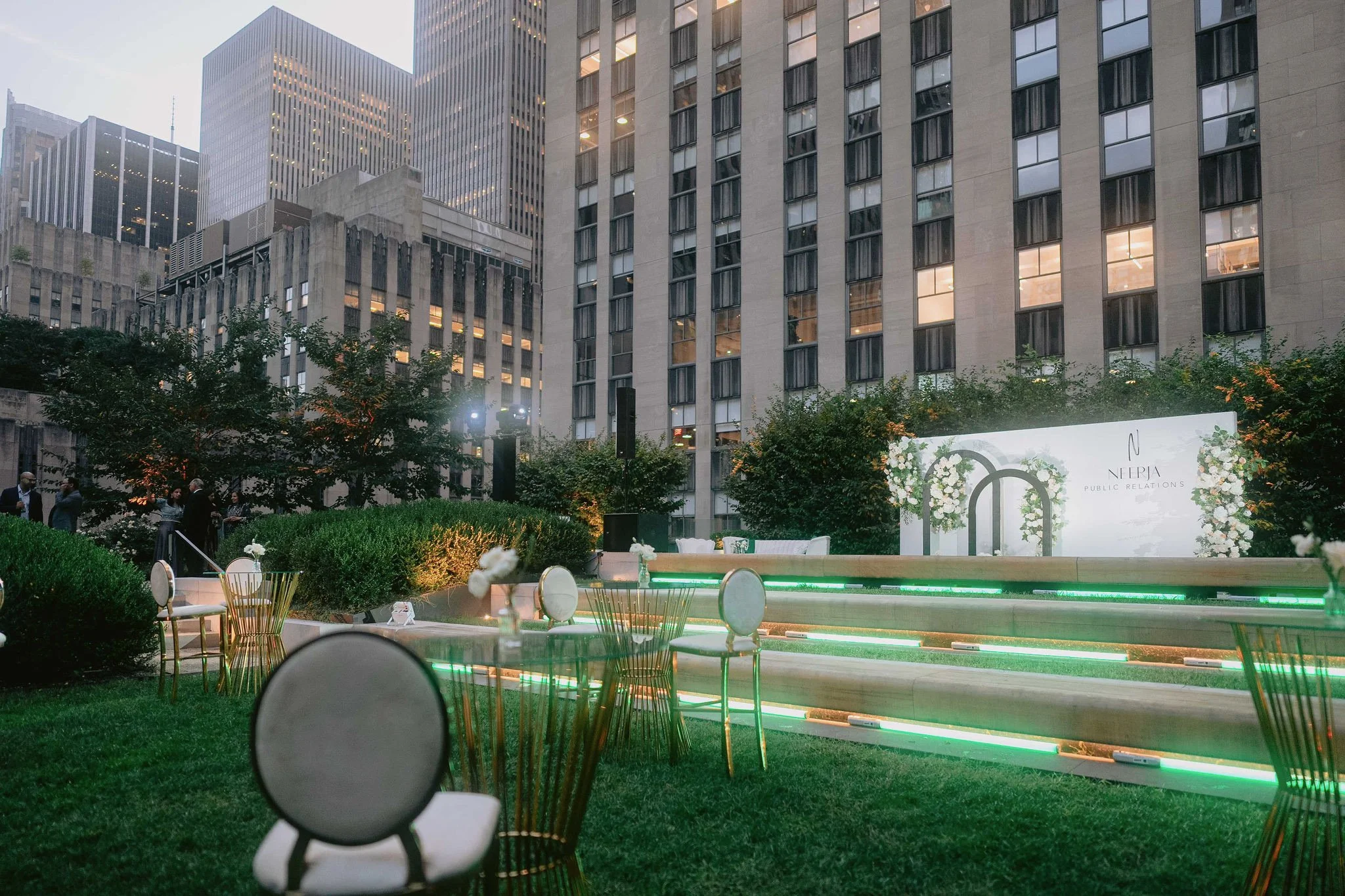 An outdoor event space in an urban setting with tables and chairs, greenery, and a lit stage area. Tall office buildings are visible in the background.
