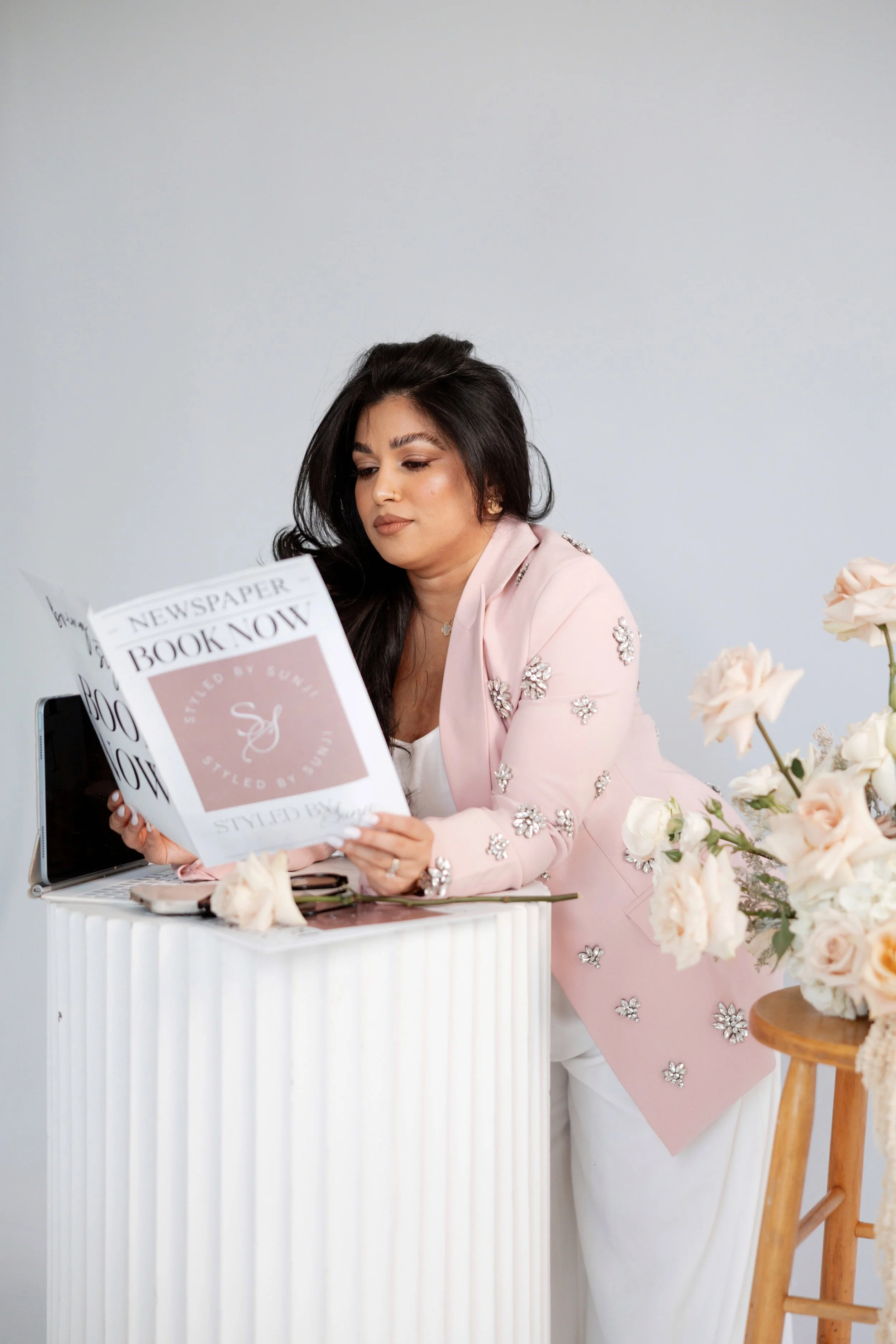 A woman with long dark hair, wearing a pink jacket adorned with decorative flowers, is reading a newspaper titled 'Book Now.' She is standing next to a white table with a vase of light pink roses, a smartphone, and a set of glasses, with a plain whit