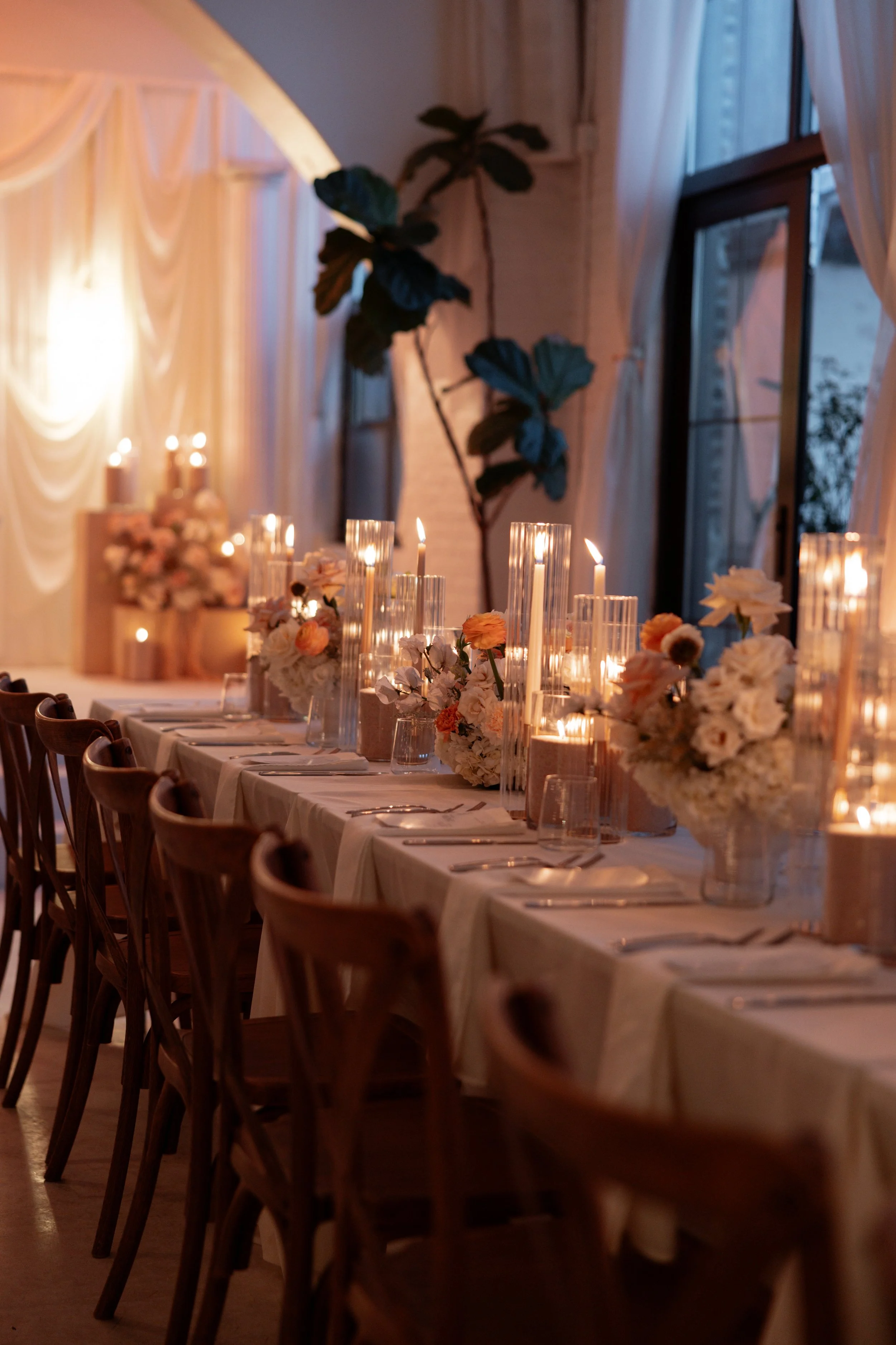 Elegant dinner table setup with white tablecloth, floral centerpieces, and tall glass candleholders with lit candles, inside a warmly lit room with draped curtains and large window.