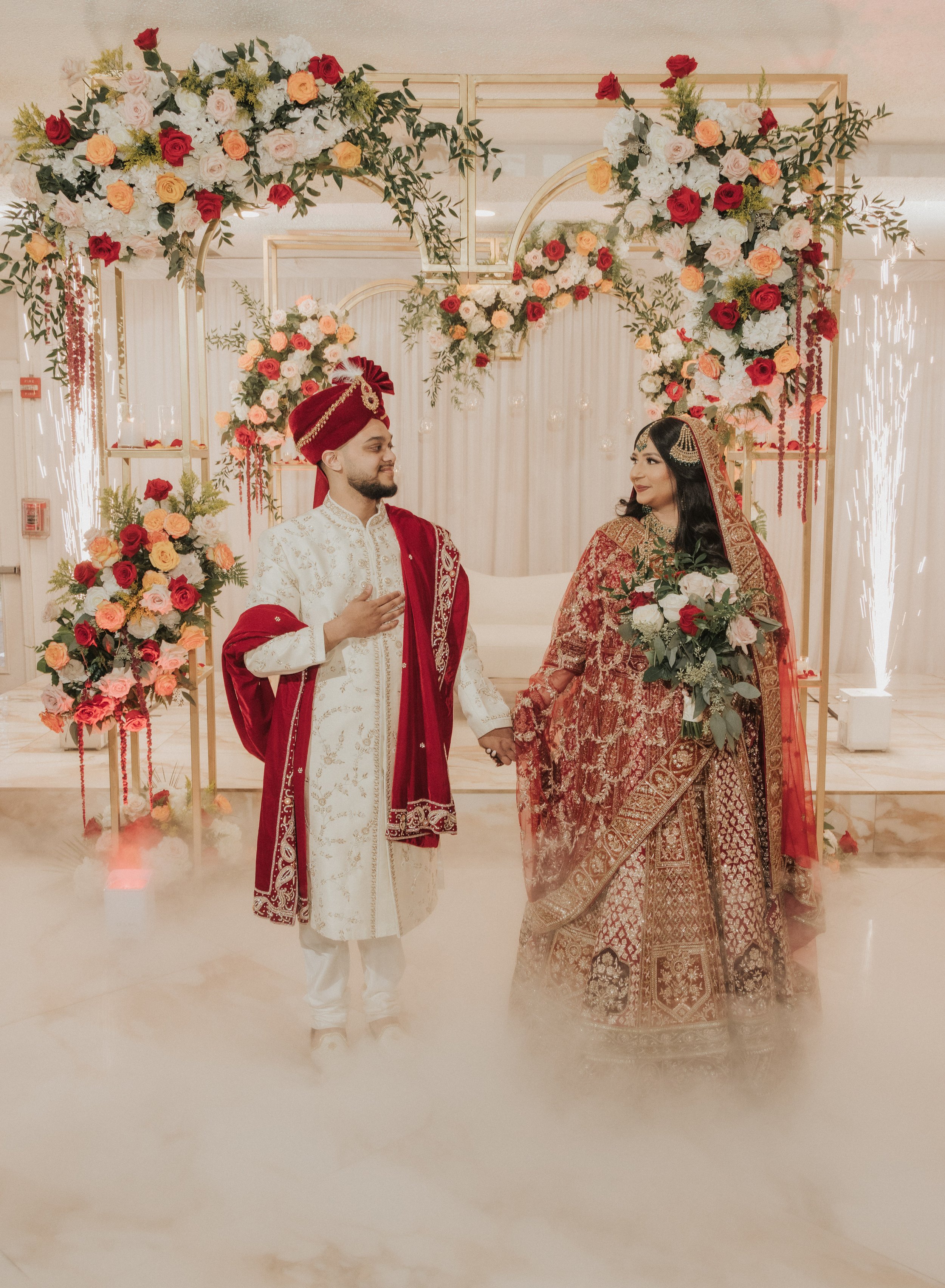 A bride and groom dressed in traditional Indian wedding attire holding hands in front of a decorated floral backdrop with arches and hanging flowers.