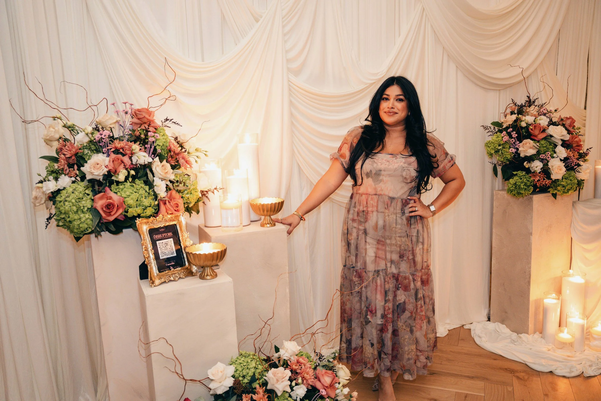 Woman standing in front of floral arrangements and white draped backdrop at an event.