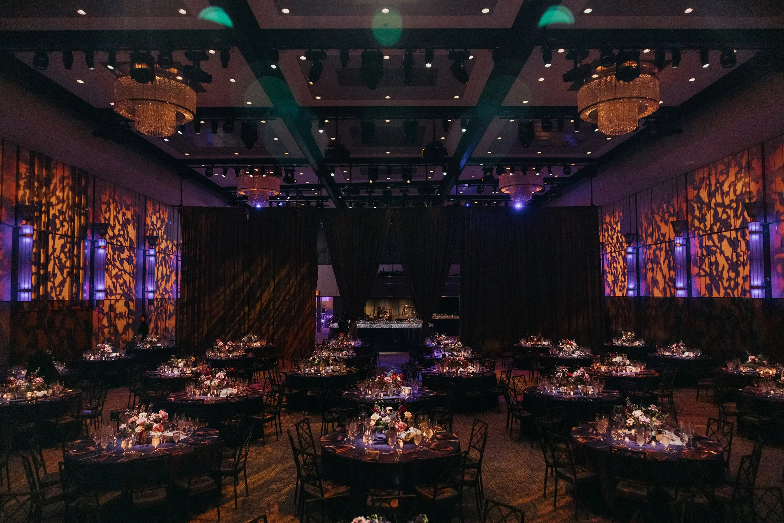 Elegant banquet hall with tall ceilings, large chandeliers, and decorated walls illuminated in purple and orange patterns. Round tables set with floral centerpieces, candles, and formal place settings are arranged across the room, suggesting a specia