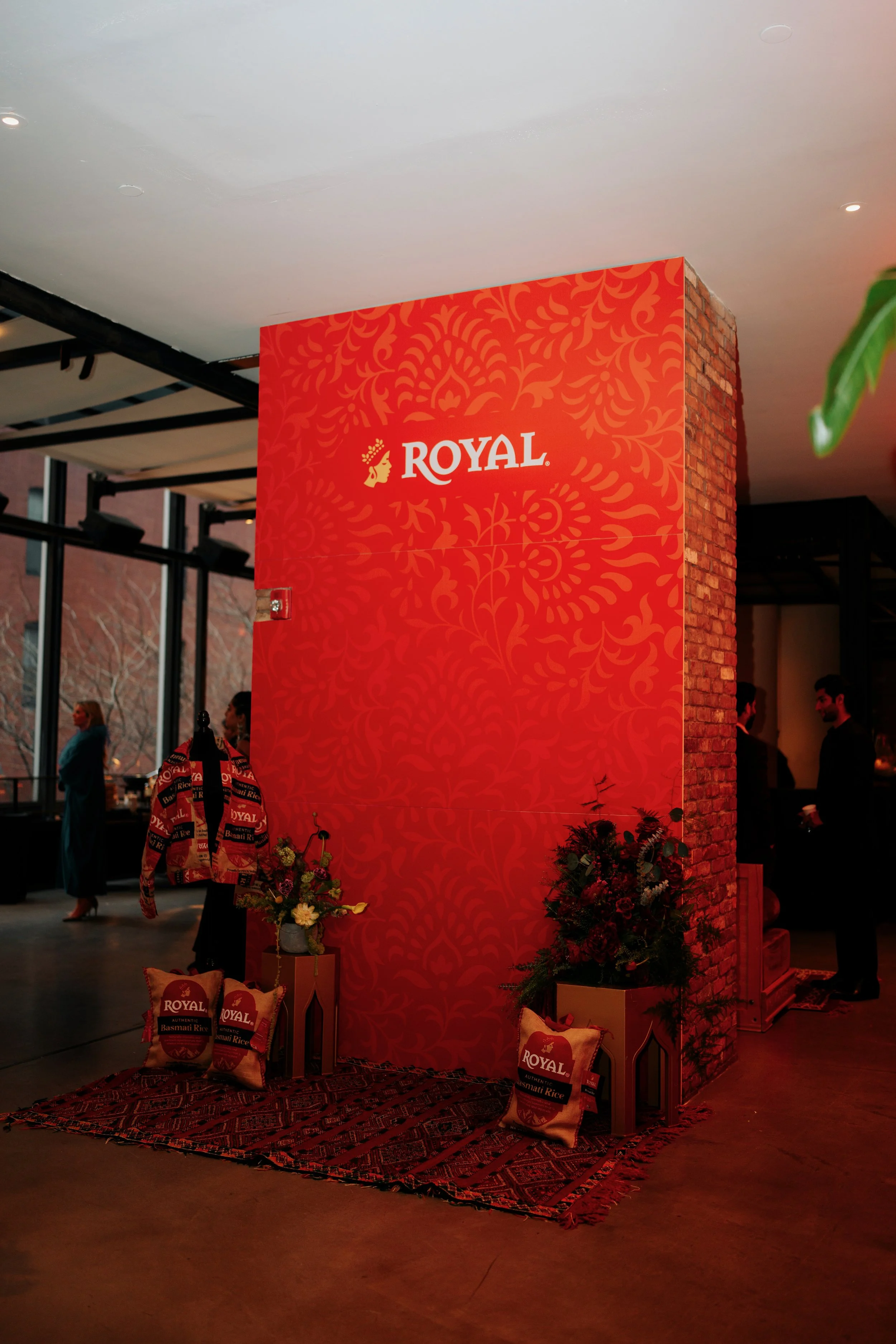 Indoor event featuring a red backdrop with the brand name 'Royal' and a logo of a woman's profile. There are floral arrangements, pillows with rice brand logos, and a traditional patterned rug on the floor. Several people are visible in the backgroun