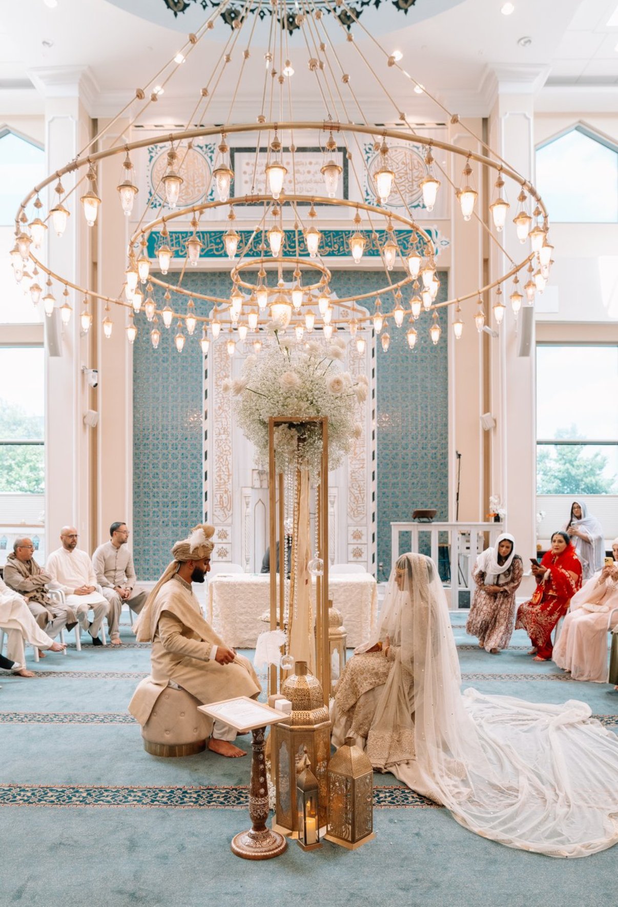 A wedding ceremony in a beautifully decorated indoor space with a blue and white theme, featuring a large floral centerpiece, a gold chandelier, and people dressed in traditional attire, including the bride in a white gown with a veil and the groom i