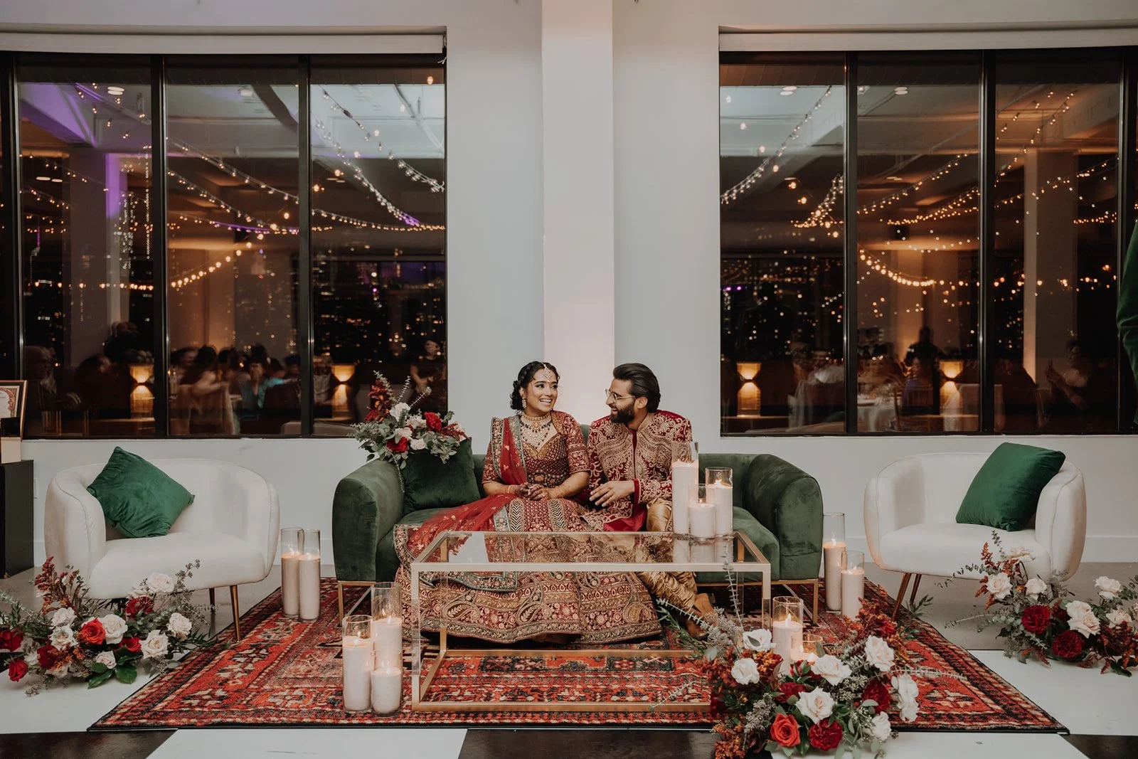 A couple dressed in traditional Indian attire sitting on a green velvet sofa at a festive event, with candles and floral arrangements on a glass table, large windows revealing a cityscape at night, and guests in the background.