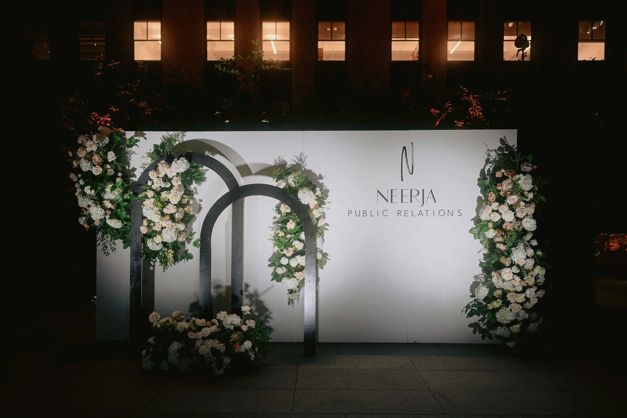A floral backdrop with white and blush roses, greenery, and decorative black arches in front, with a sign displaying 'NEERJA PUBLIC RELATIONS' in black text on a white background, set at night.