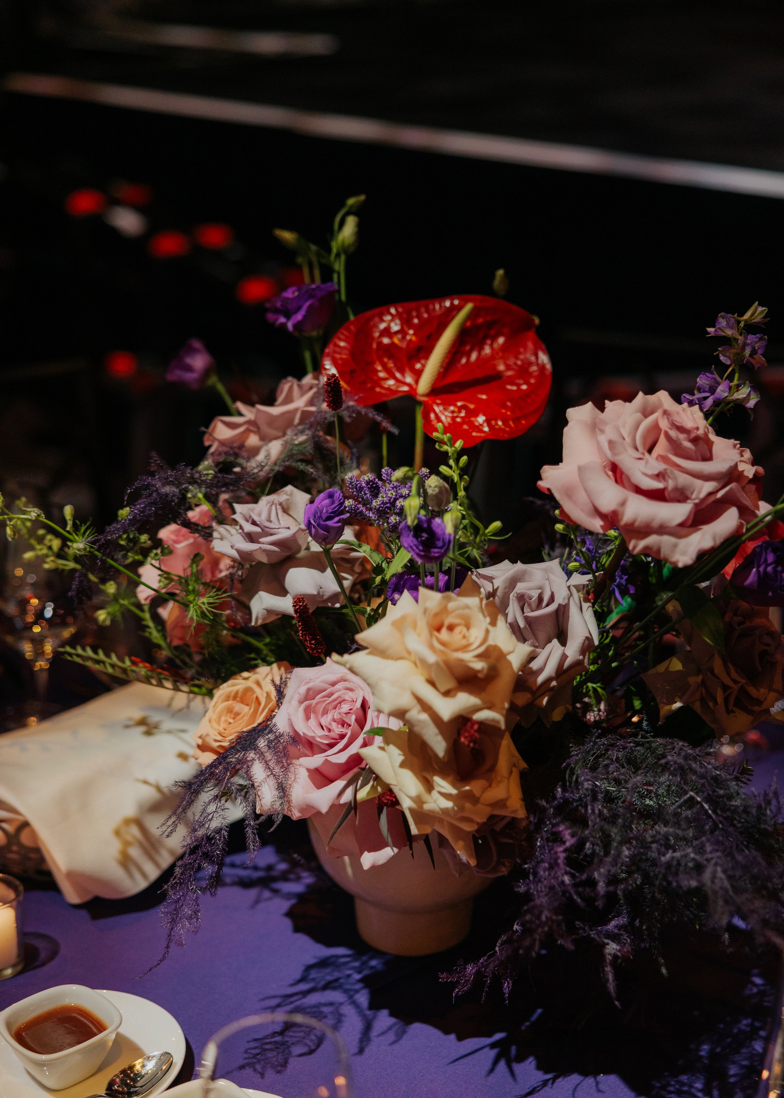A floral arrangement of pink roses, purple flowers, and a red anthurium in a vase, with a small bowl of sauce and spoon in the foreground on a purple tablecloth.