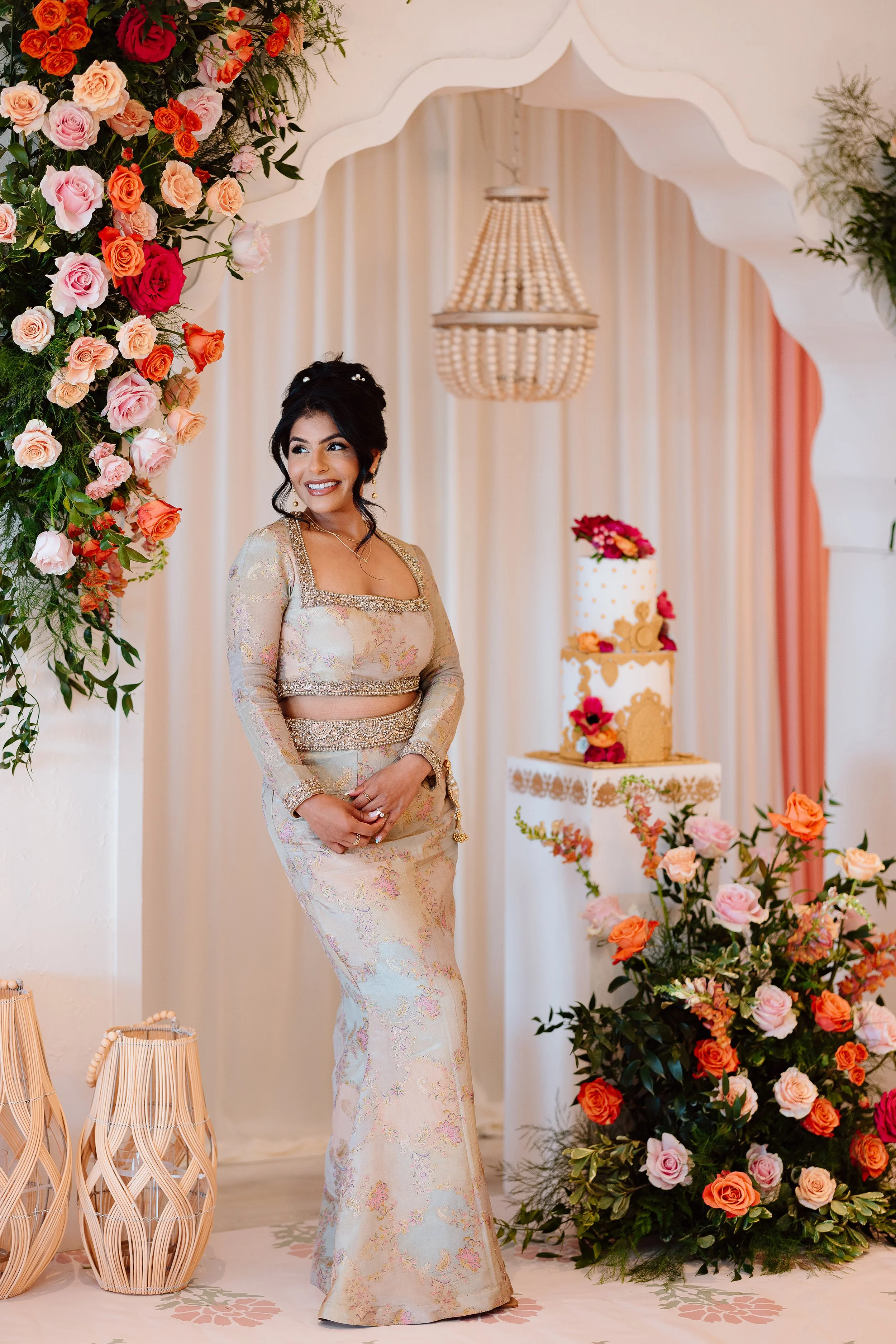 A woman wearing a traditional, elegant dress stands amidst floral decorations and a wedding cake, smiling during a celebration or special event.