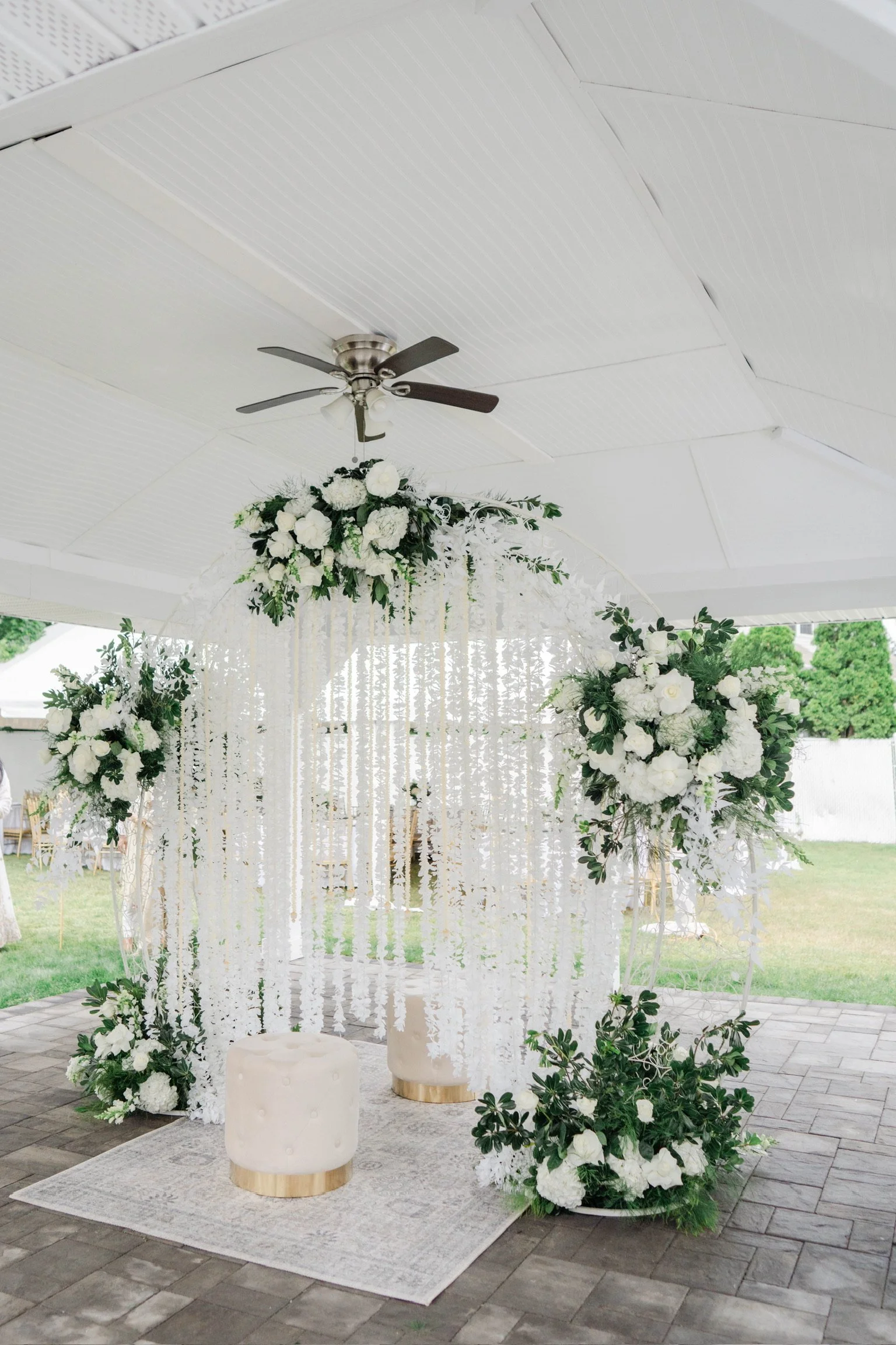 White floral wedding arch decorated with white flowers and green foliage, set under a white pavilion with hanging ribbons and a patterned white rug on the ground.