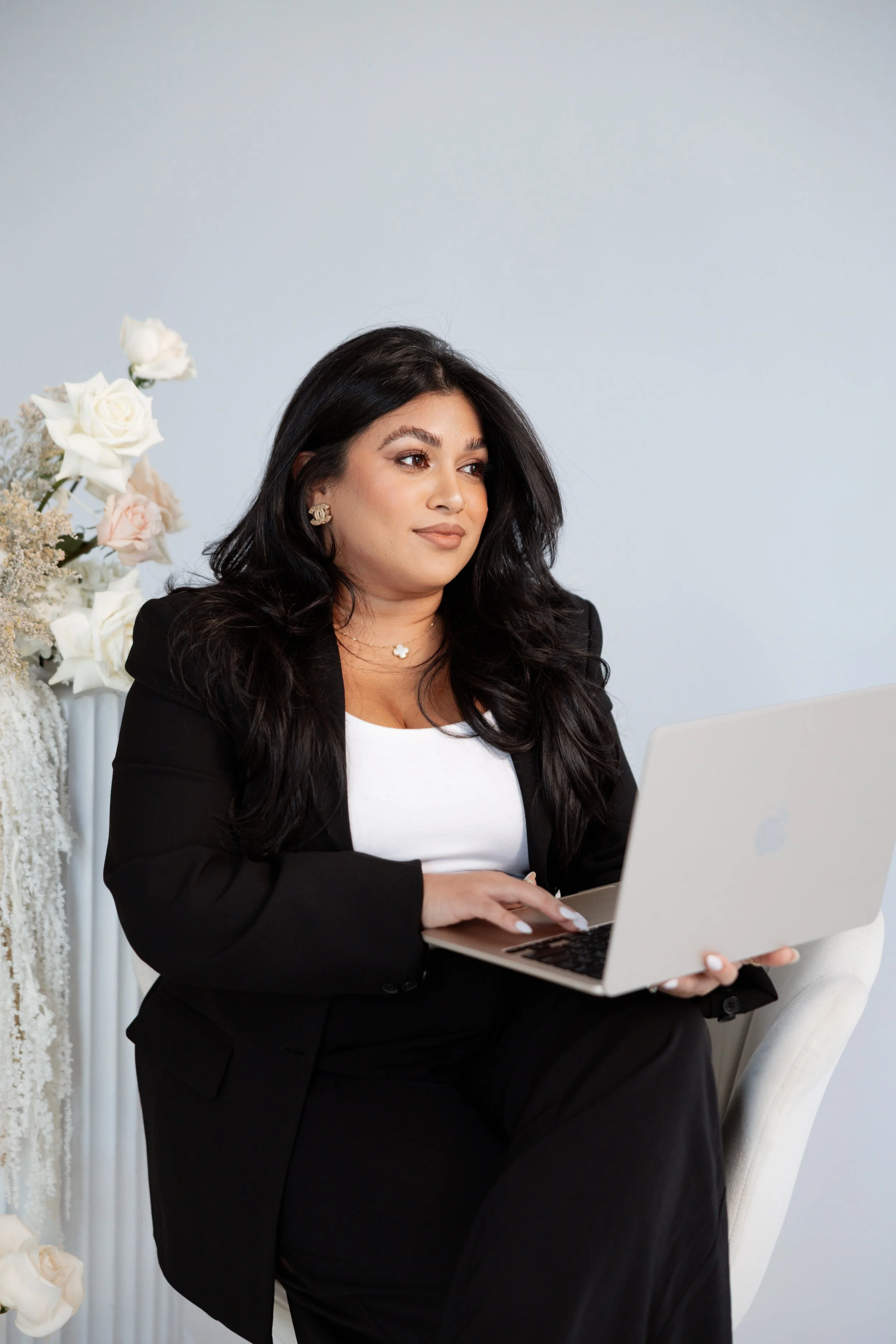A woman with long black hair, wearing a black blazer and white top, sitting on a white chair, working on a silver laptop, with a floral arrangement including white and pink roses beside her, in a bright room with light-colored walls.