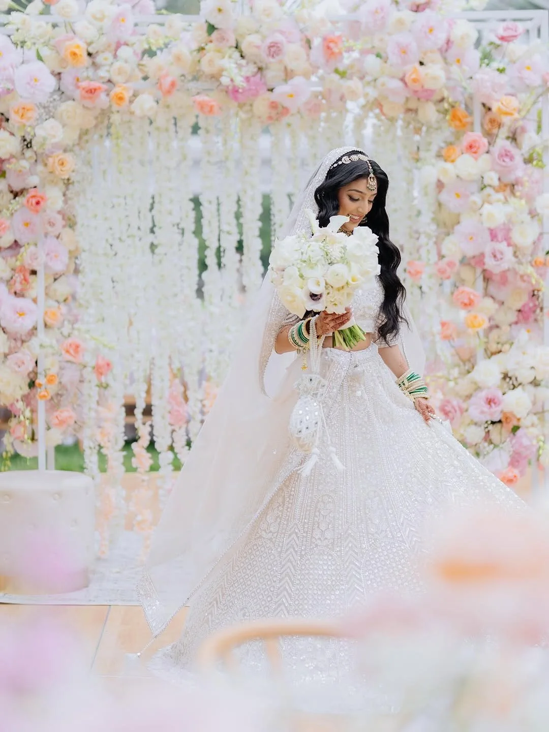 Bride in white wedding dress holding a bouquet of white flowers at a floral wedding arch.