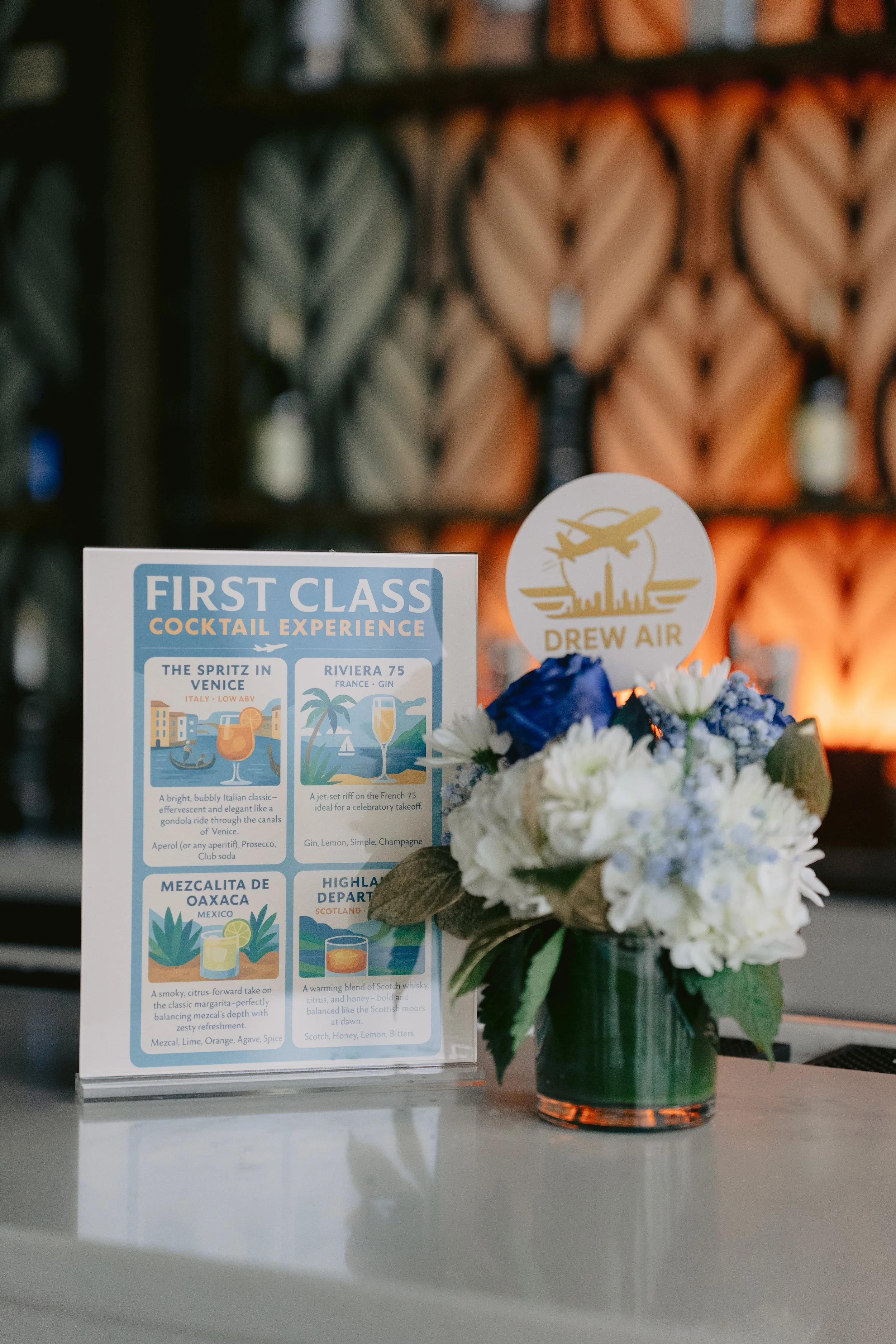 A floral arrangement with white and purple flowers next to a drink menu titled 'First Class Cocktail Experience' and a round sign with an airplane logo and 'Drew Air' on it. The background features a wall with hanging glasses or bottles.