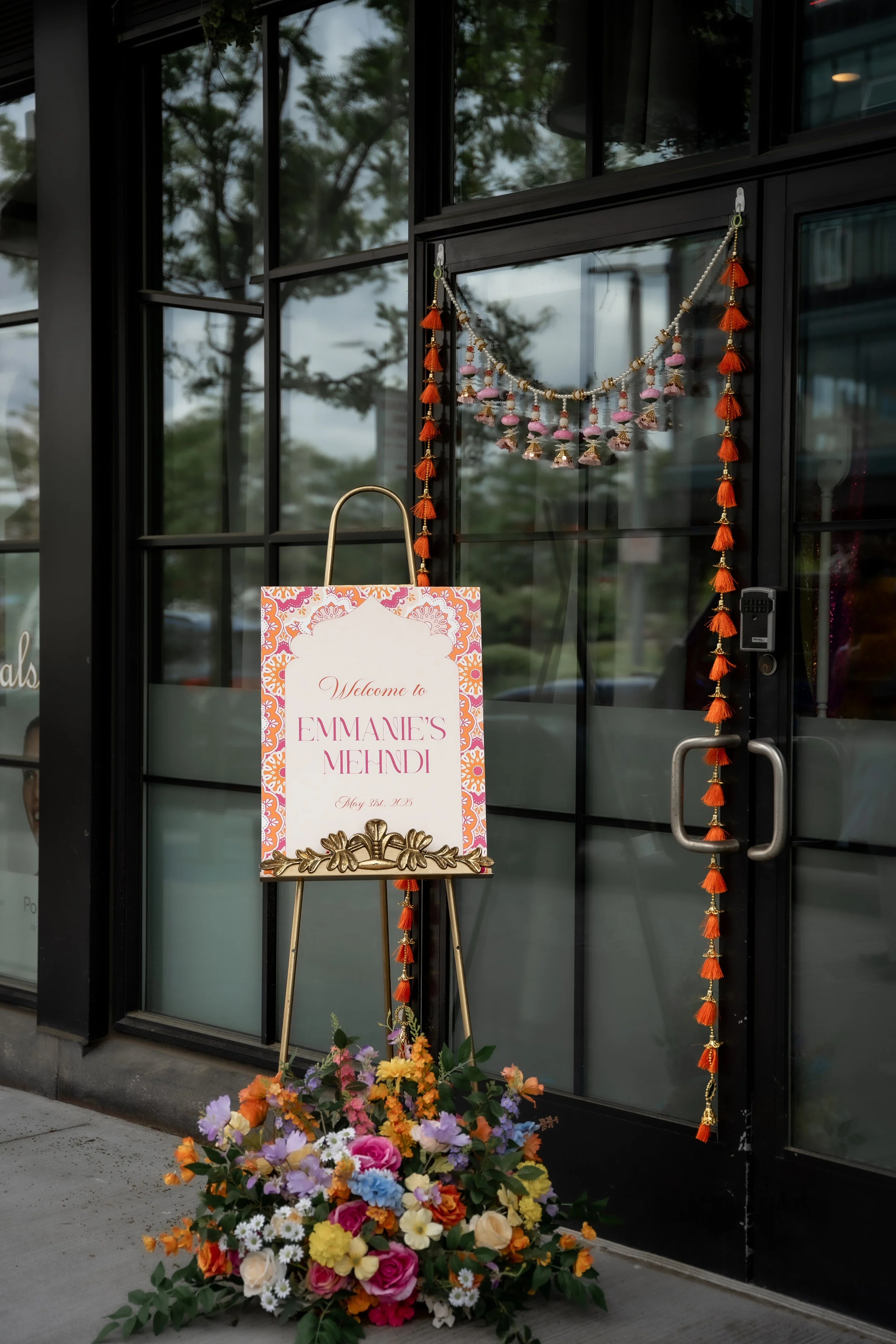 A decorative sign reading 'Welcome to Emmanee's Mehndi' is placed on a gold easel outside a building. There is a large, colorful flower arrangement at the base of the sign. The building has black-framed glass doors decorated with hanging orange tassels and strings of beads. Reflections of trees can be seen in the glass.