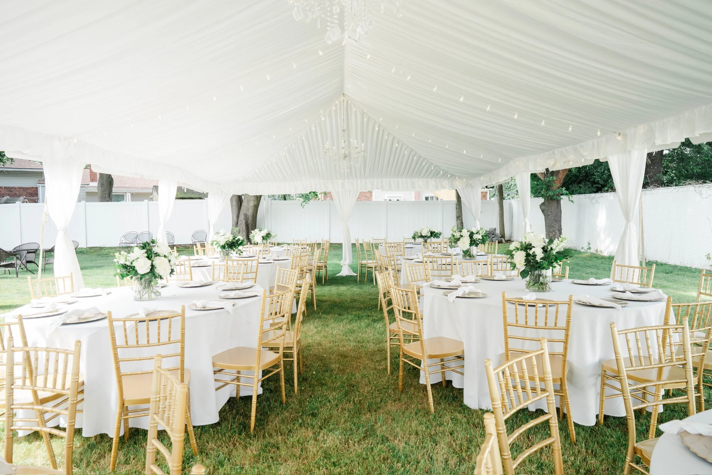 An outdoor event setup with white round tables covered with white tablecloths, each decorated with floral centerpieces, and surrounded by light-colored wooden chairs. A white tent with draped fabric and hanging lights covers the area, with a grassy y